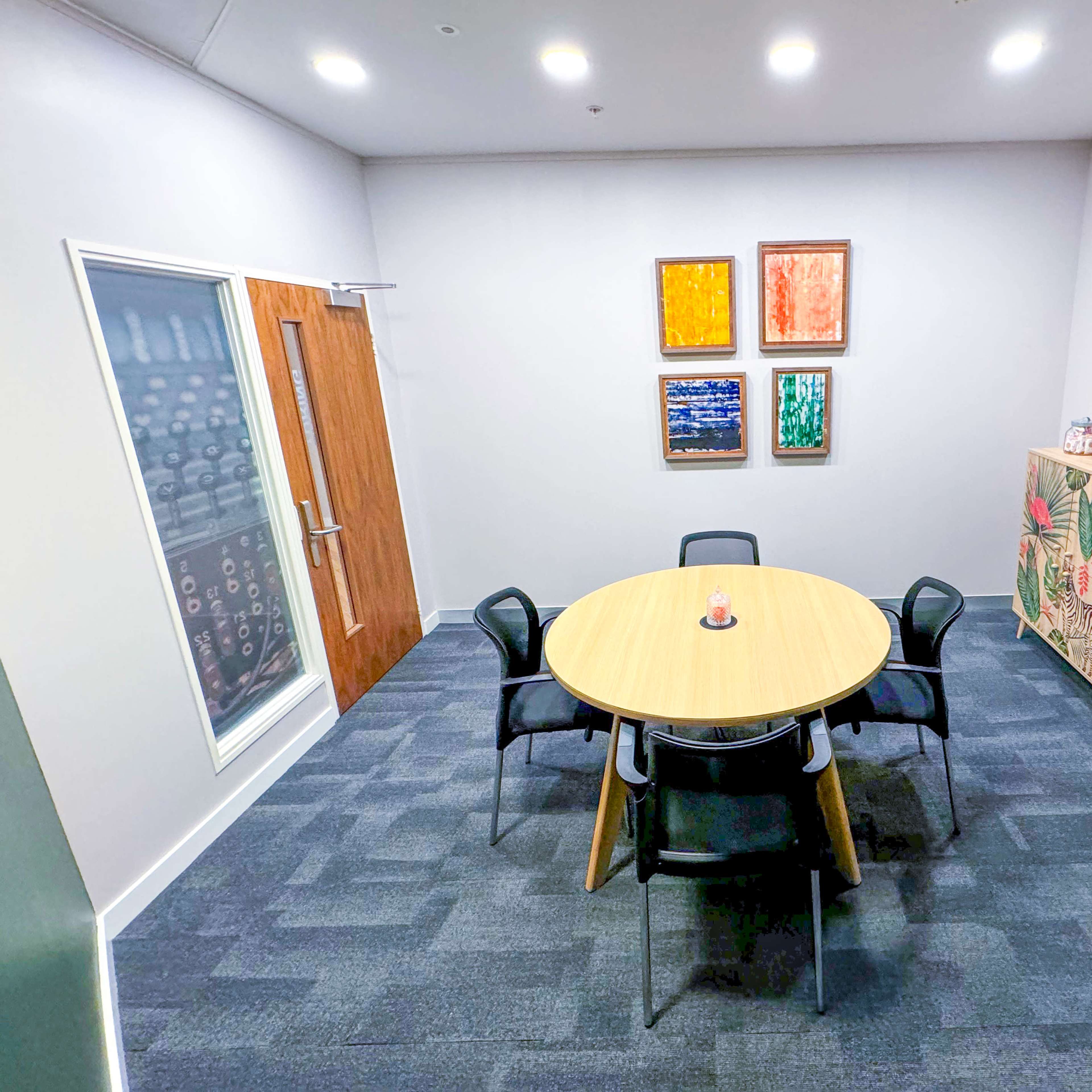 A small meeting room features a round wooden table surrounded by six chairs, with colorful artwork on the walls and a cabinet in the corner.