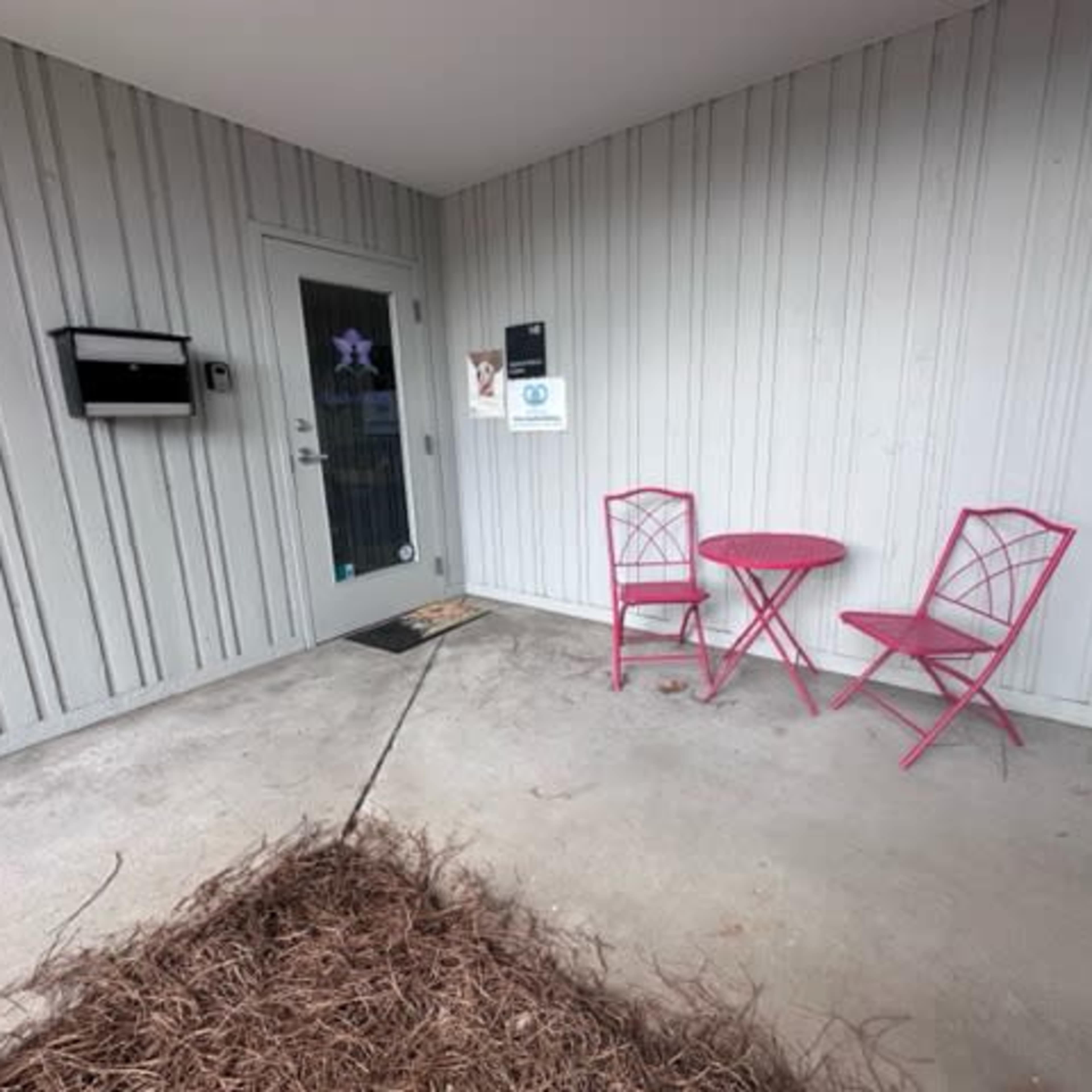 A small entrance area featuring a gray wooden wall, a door with a window, a mailbox, and a pink table with two matching chairs.