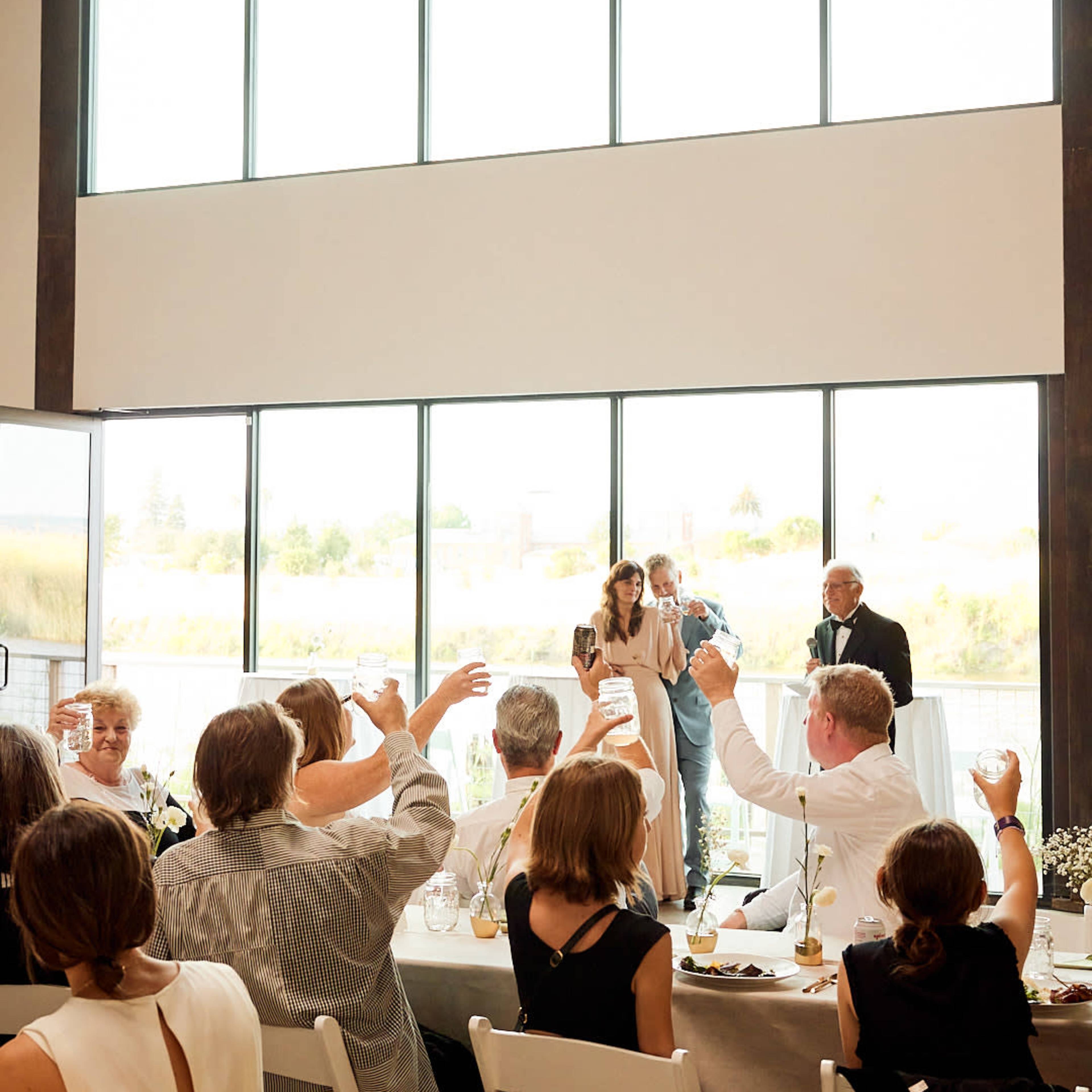 A couple stands together while guests raise their glasses for a toast in a bright, spacious indoor venue with large windows.
