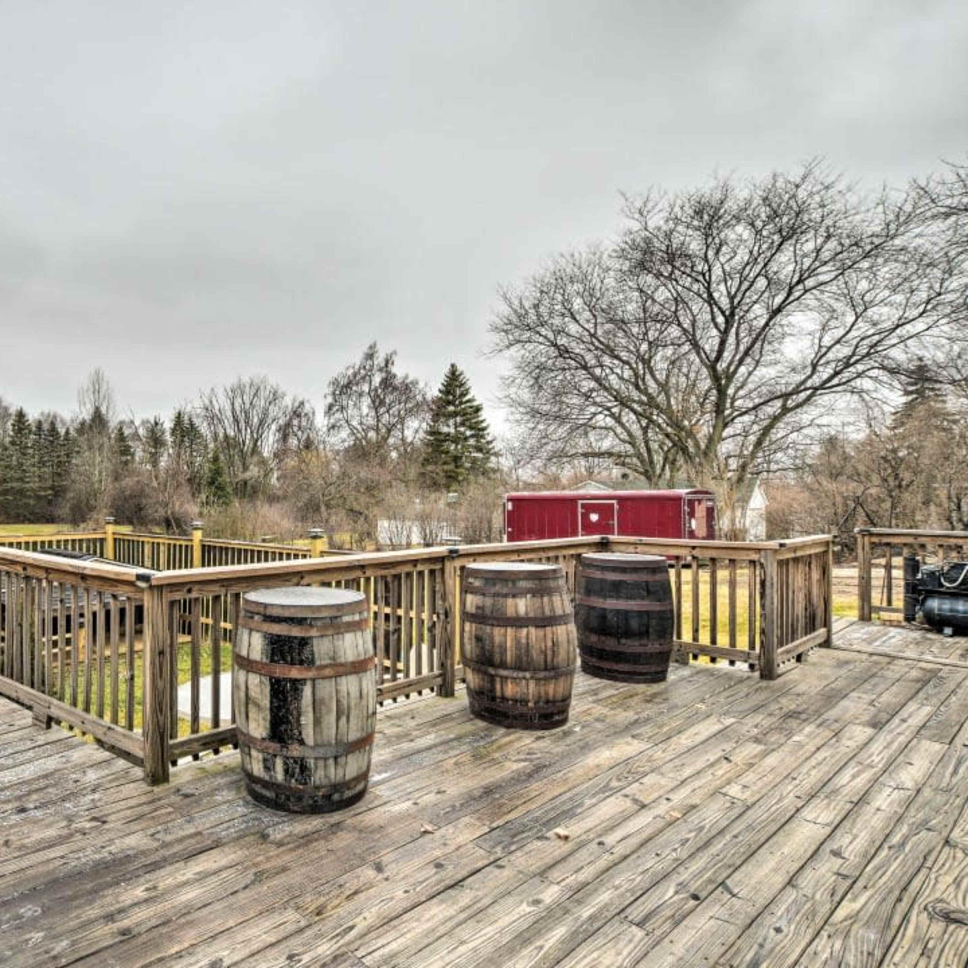 A wooden deck with three barrels placed on it, surrounded by trees and distant buildings under a cloudy sky.