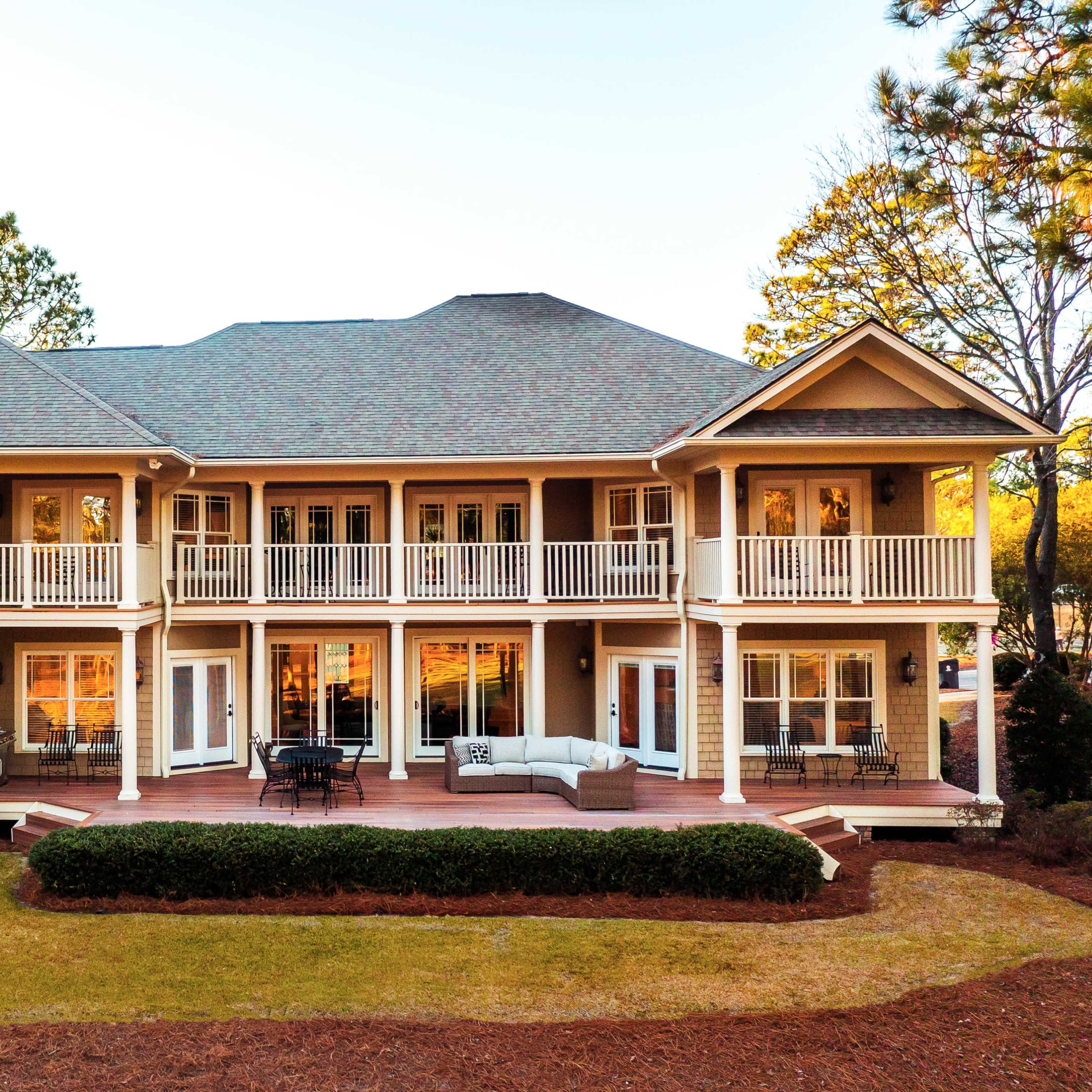 A two-story house with a large deck featuring multiple seating areas overlooks a landscaped yard.