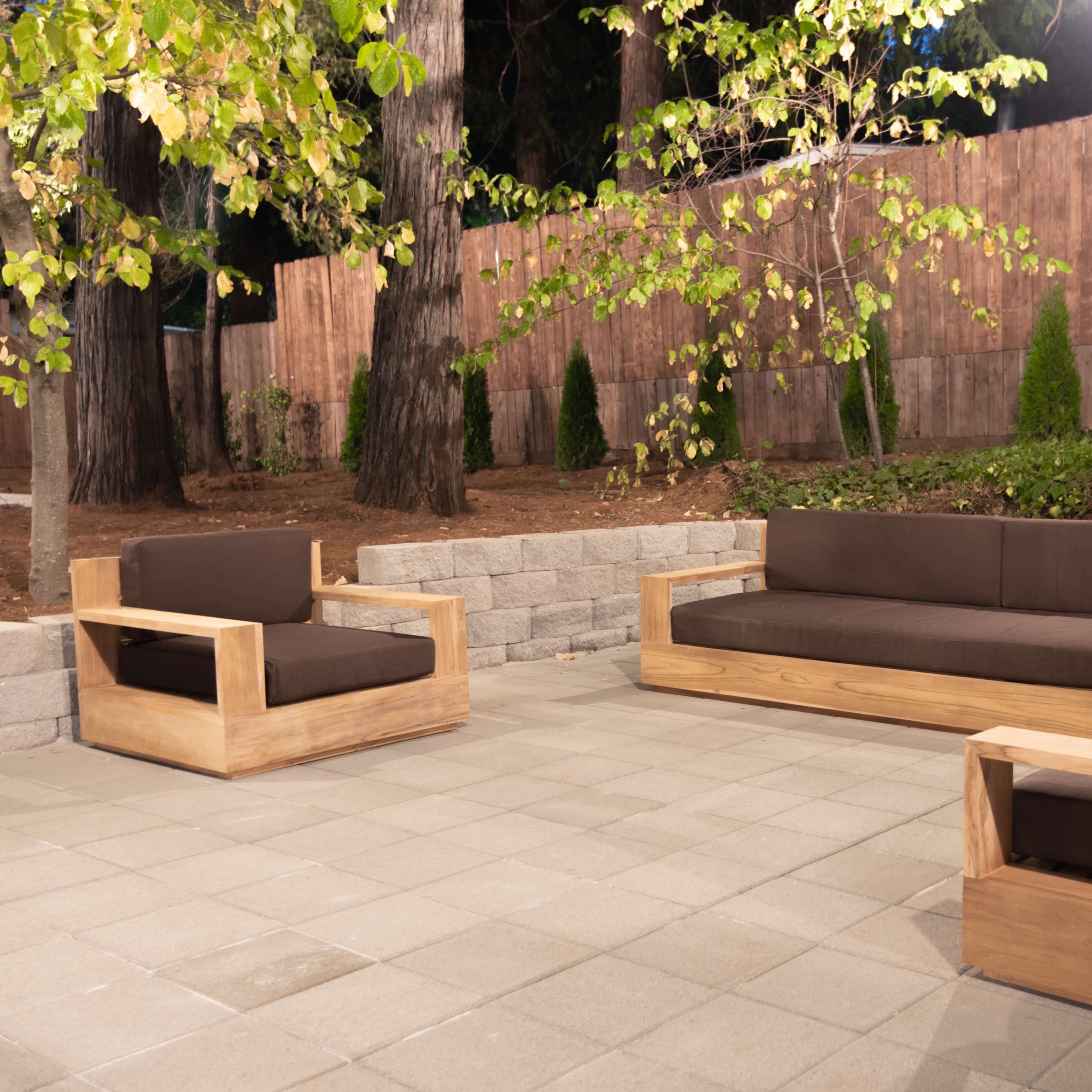 A modern outdoor seating area with wooden sofas and chairs arranged on a paved patio surrounded by trees and a wooden fence.