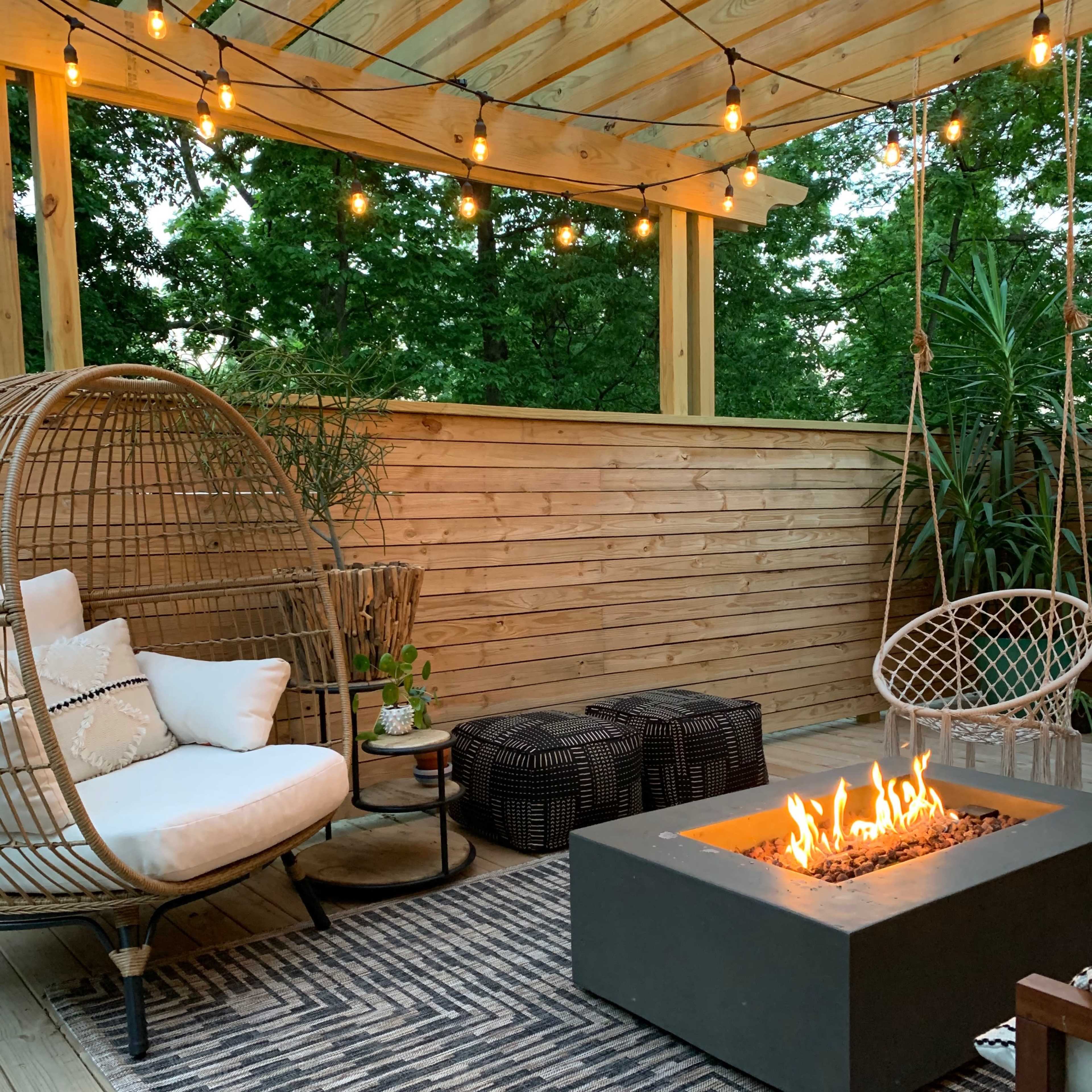 A cozy outdoor seating area with a modern fire pit surrounded by hanging lights, wicker chairs, and a patterned rug.