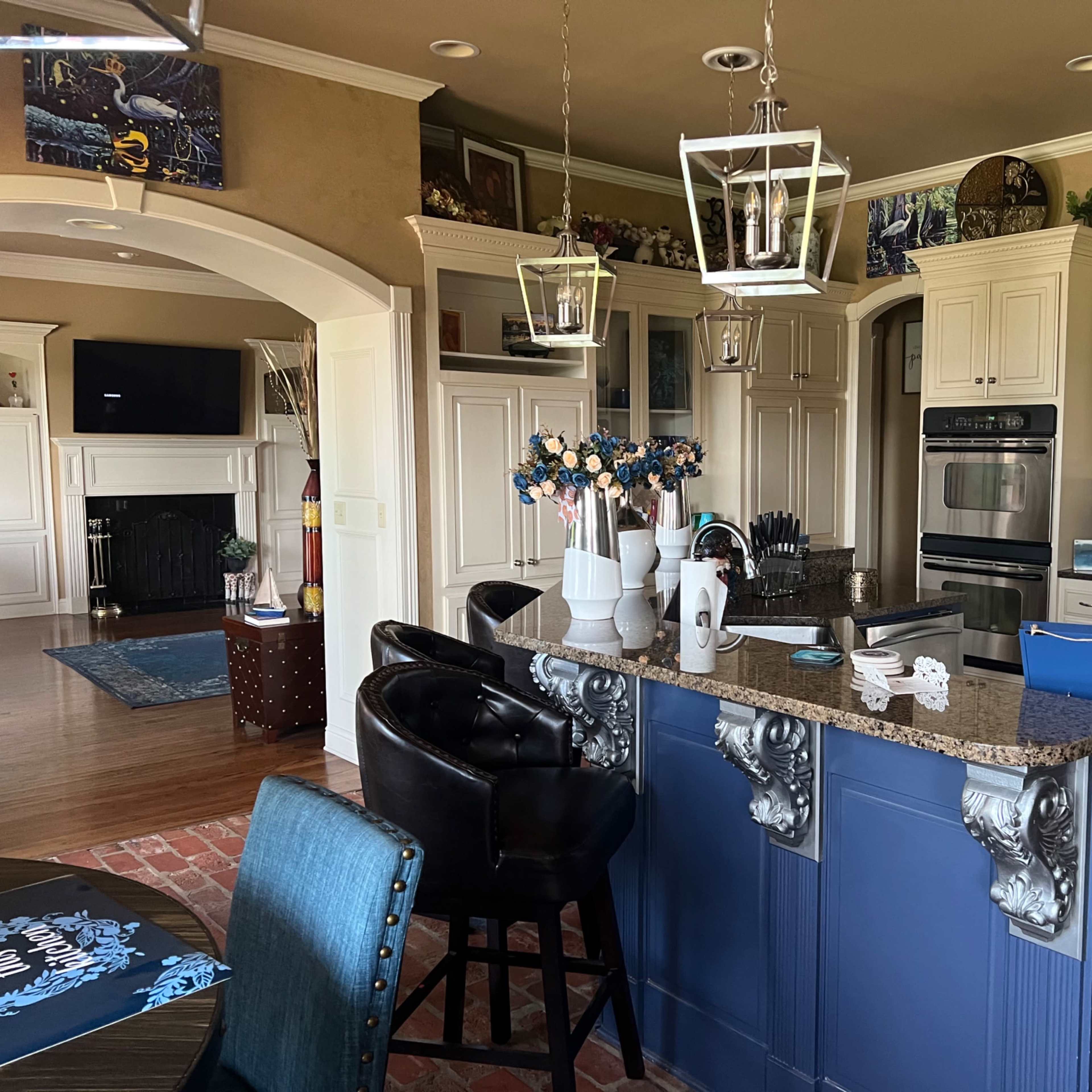 A kitchen featuring a blue island with granite countertops, black bar stools, and various decorative items on the shelves.
