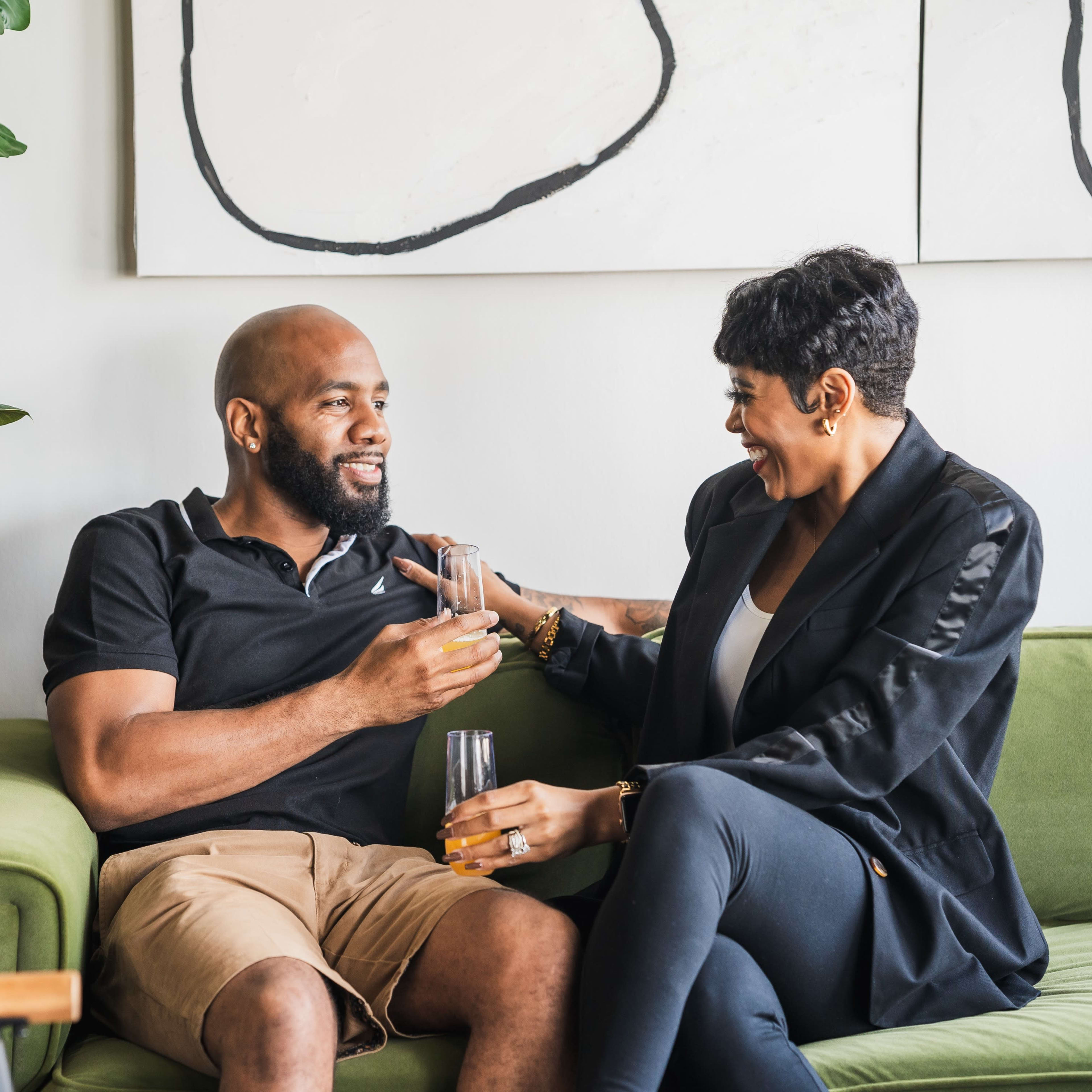 A man and woman sit together on a green sofa, laughing and enjoying drinks in a stylish living room.