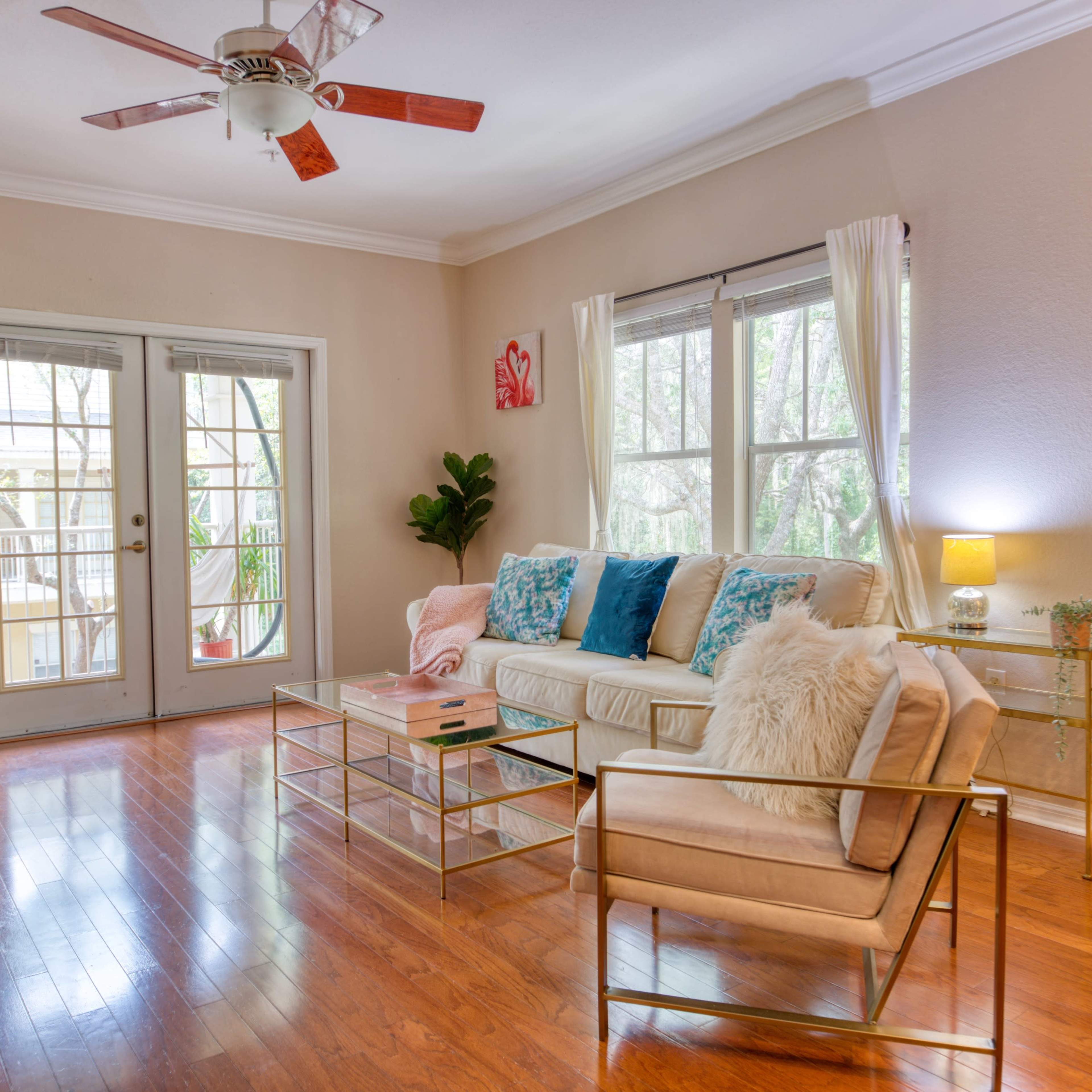The room features a light-colored sofa with decorative pillows, a glass coffee table, and large windows that open to a balcony, illuminated by natural light.
