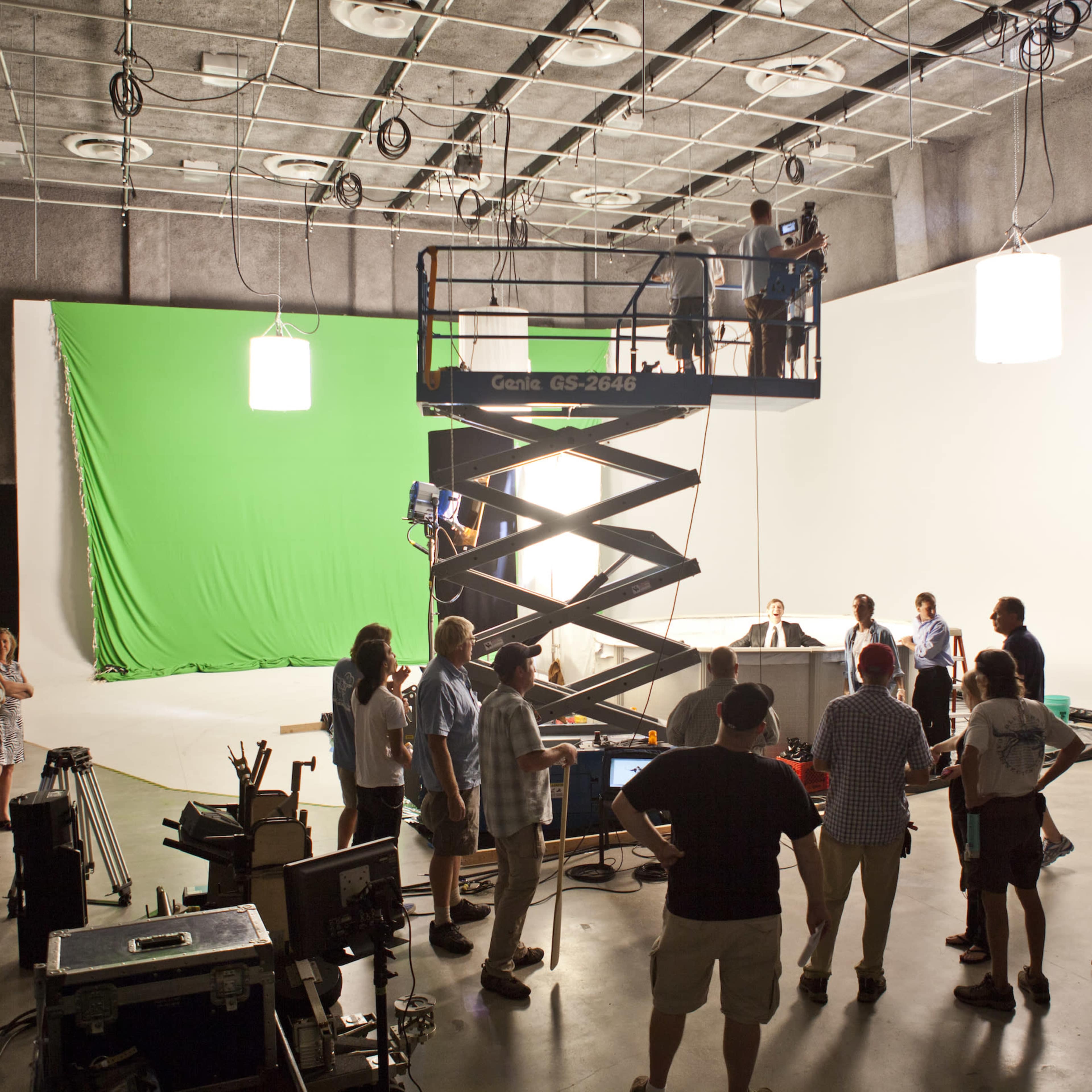 A film crew is set up in a studio, with a scissor lift in the center and a green screen backdrop on one side.