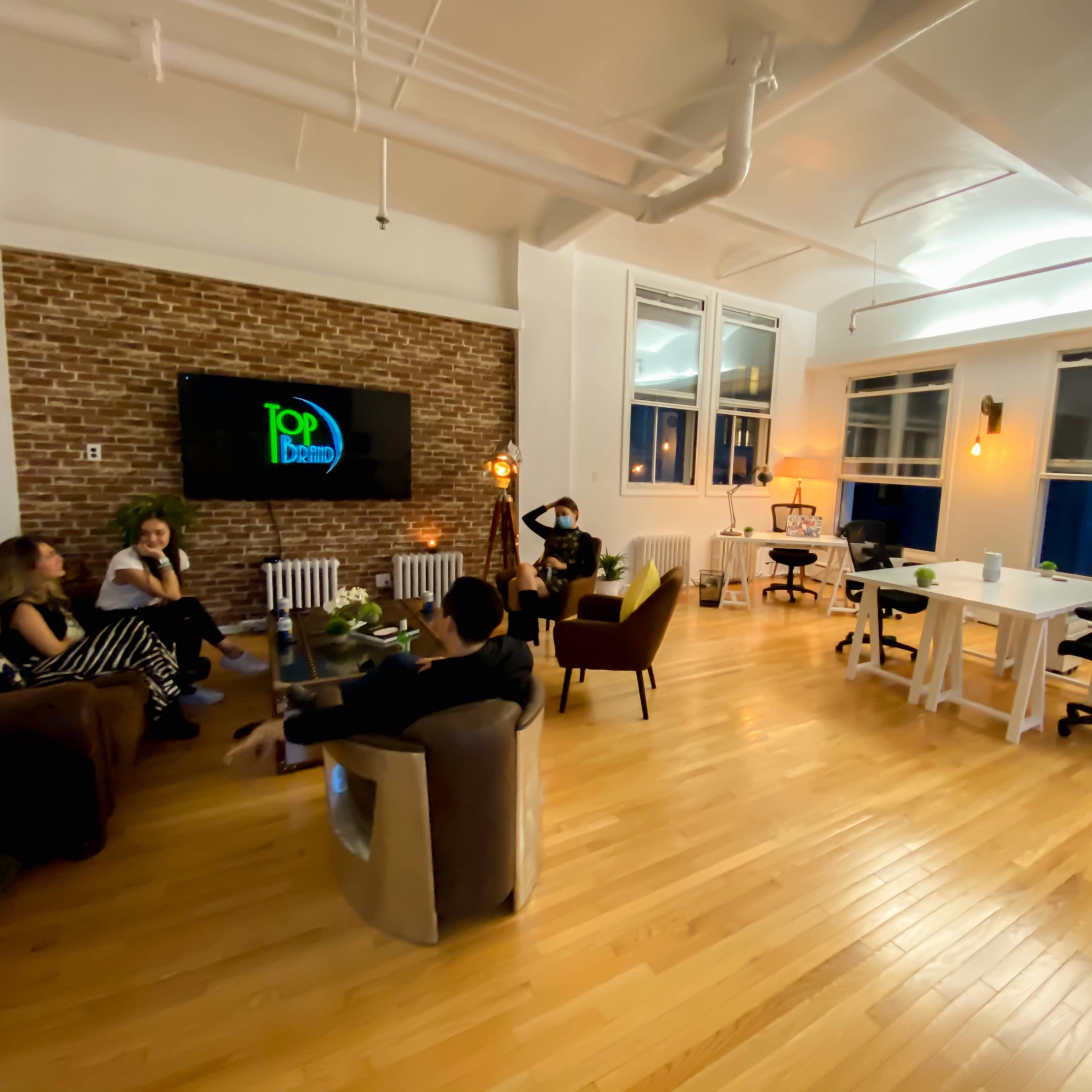 A modern co-working space features a large wooden floor, a brick accent wall, and several seating areas with people engaged in conversation.