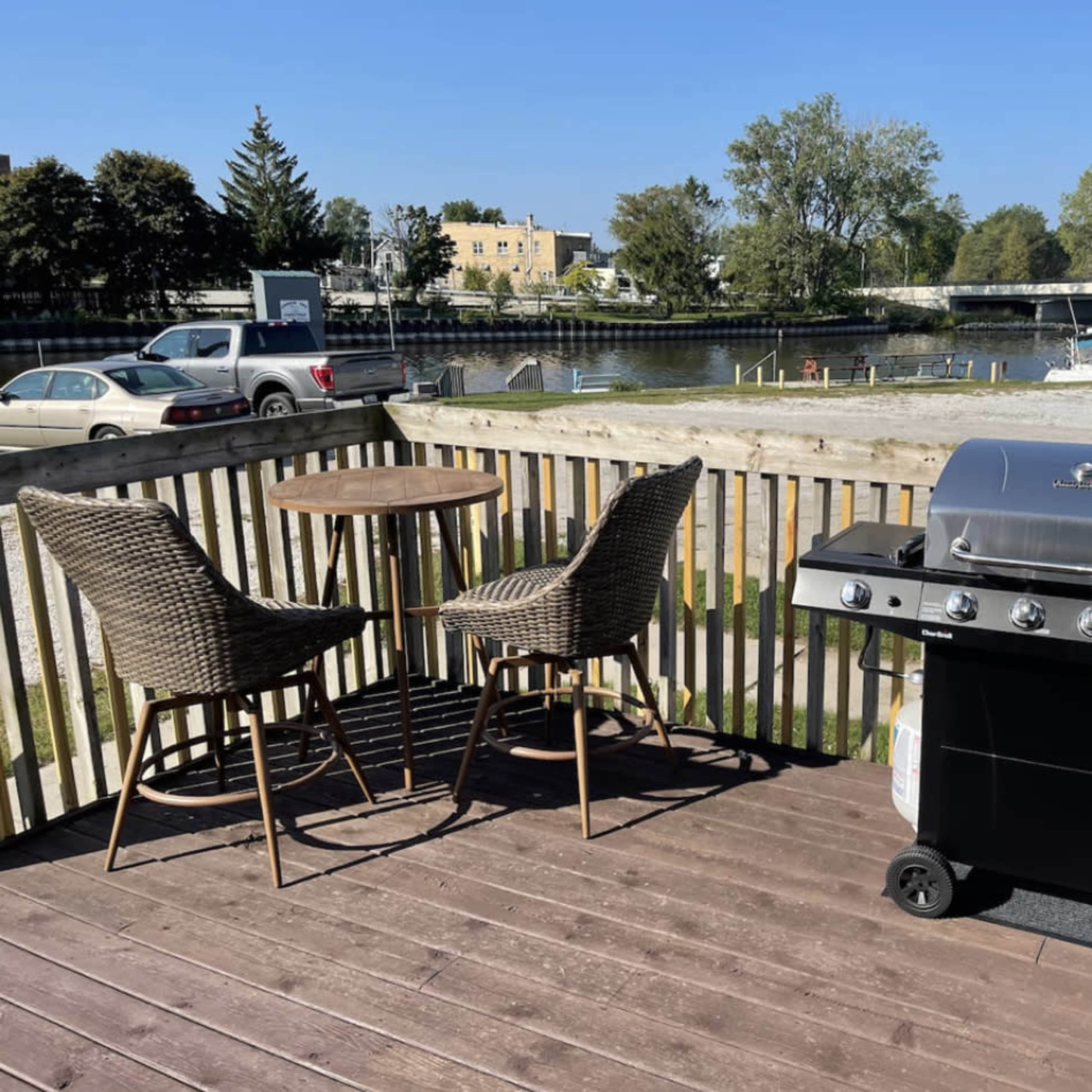 A gas grill is positioned on a wooden deck next to a round table and two wicker chairs, overlooking a waterway with boats and trees in the background.