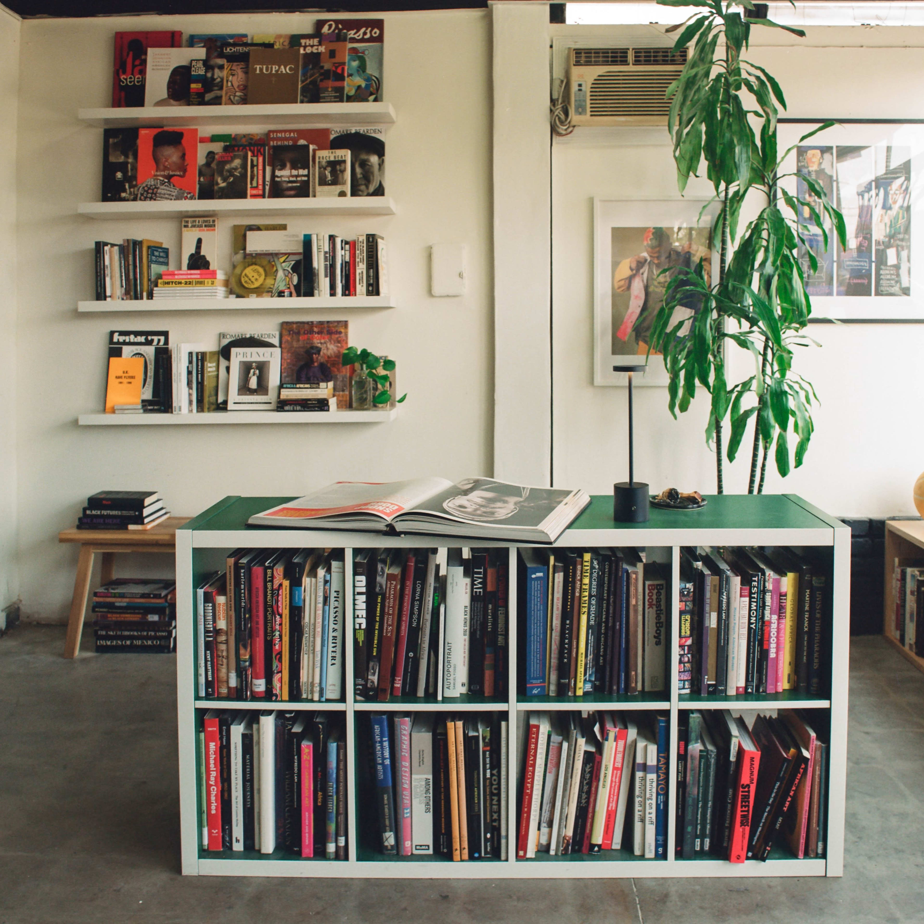 A modern reading area features a green table with bookshelves filled with various books, a potted plant, and framed artwork on the walls.