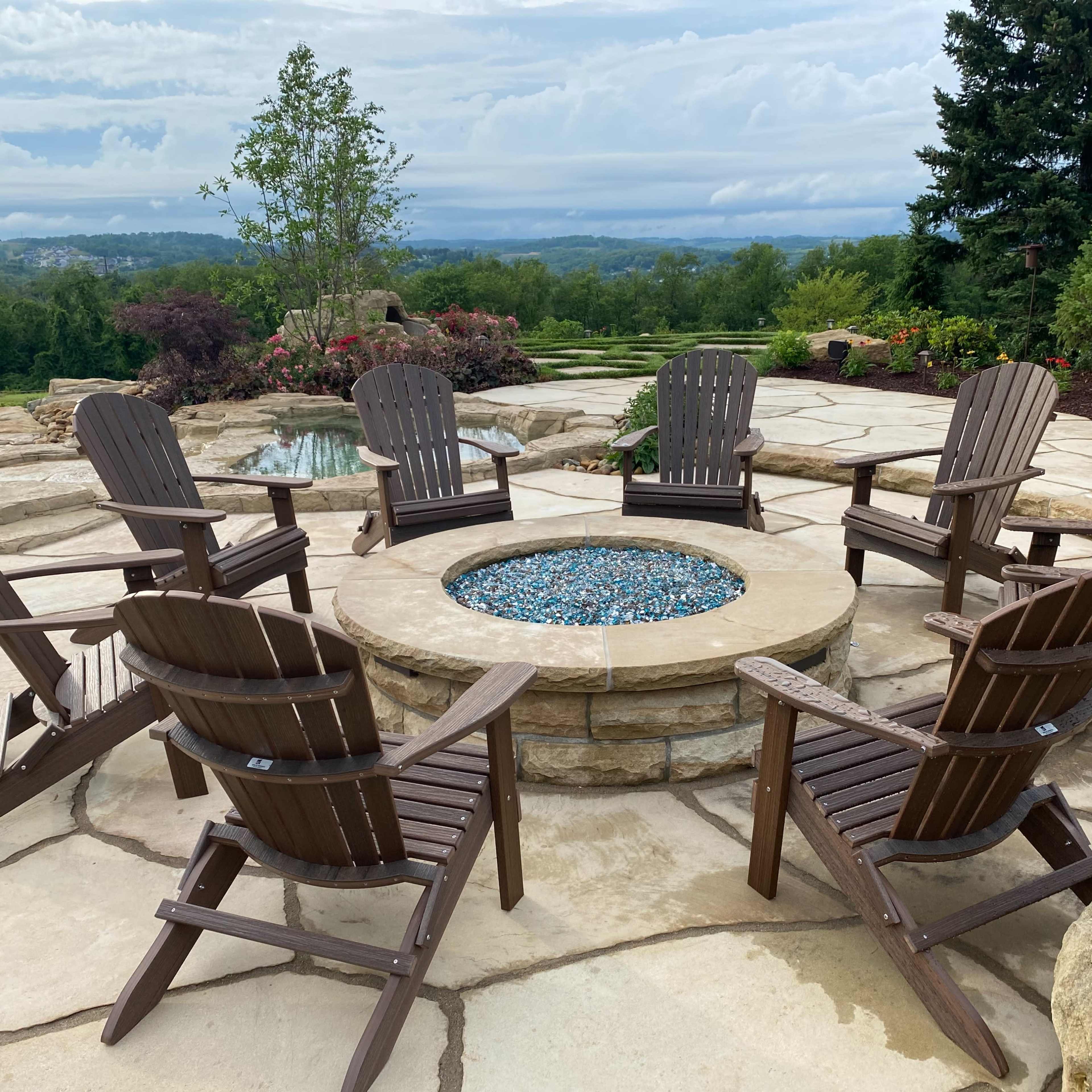A stone fire pit surrounded by eight wooden chairs sits on a paved patio overlooking a valley and lush greenery.