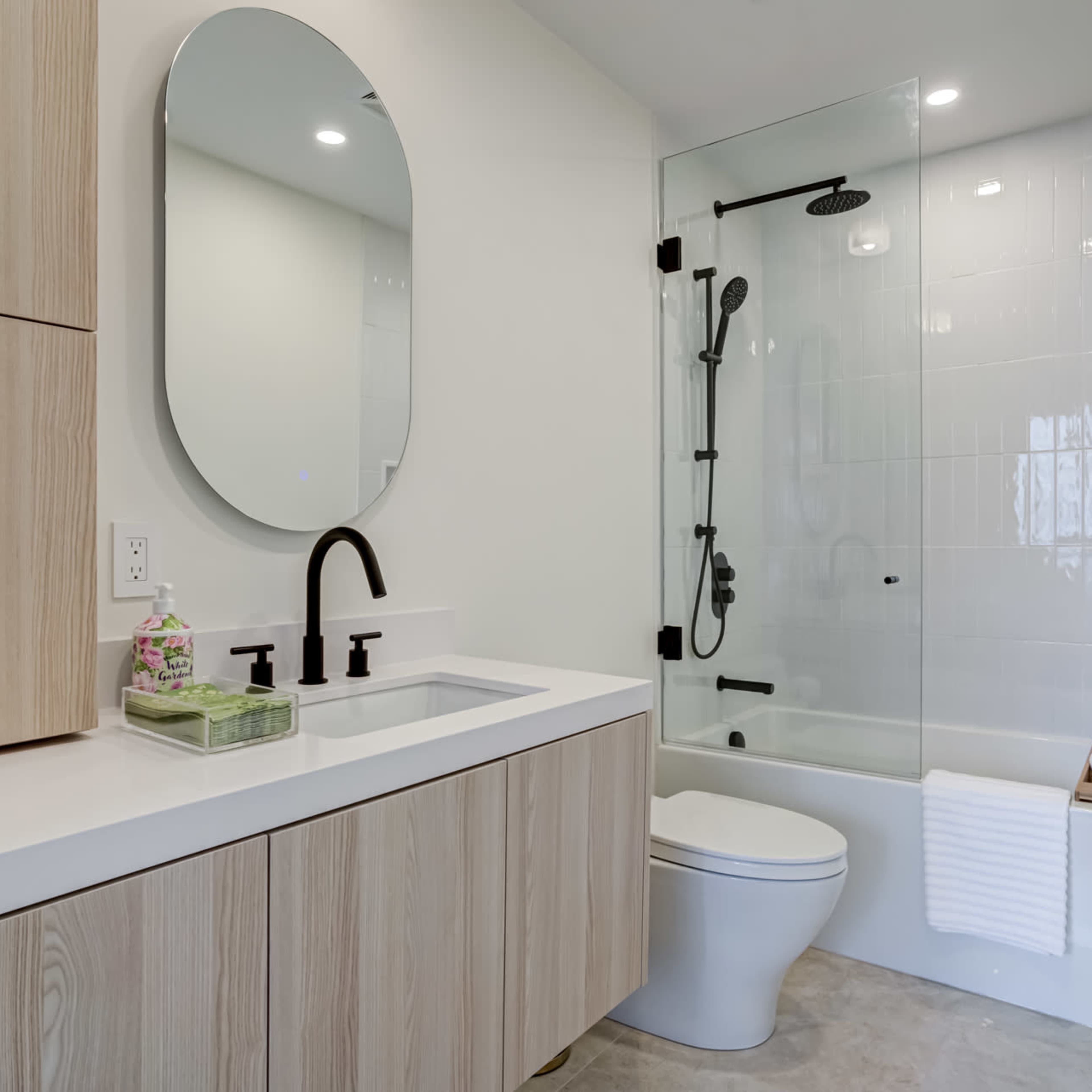 A modern bathroom featuring a minimalist design with light wood cabinetry, a round mirror, a glass shower enclosure, and a white toilet.
