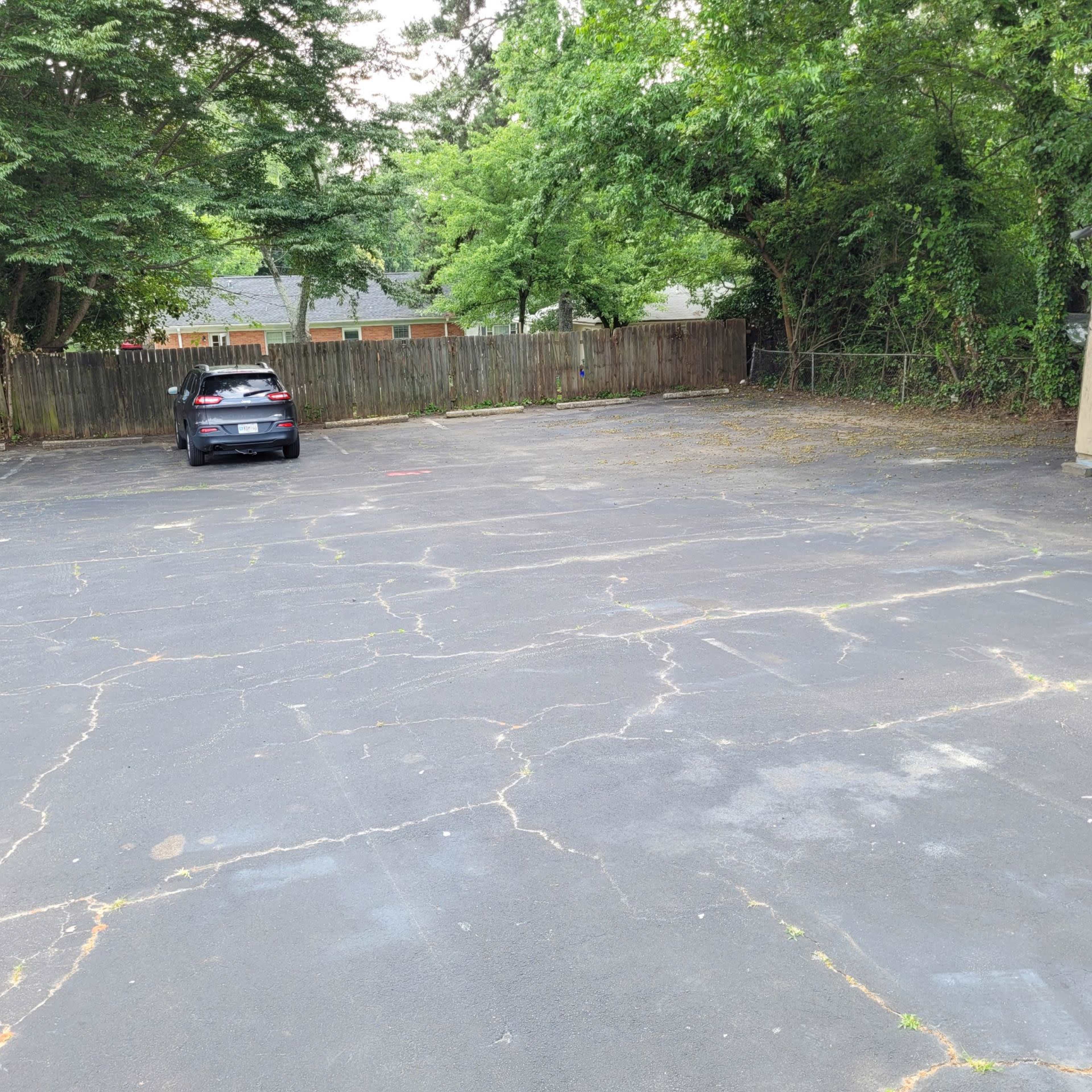 An empty parking lot with one car parked on the left side and a surrounding area of trees and a wooden fence.