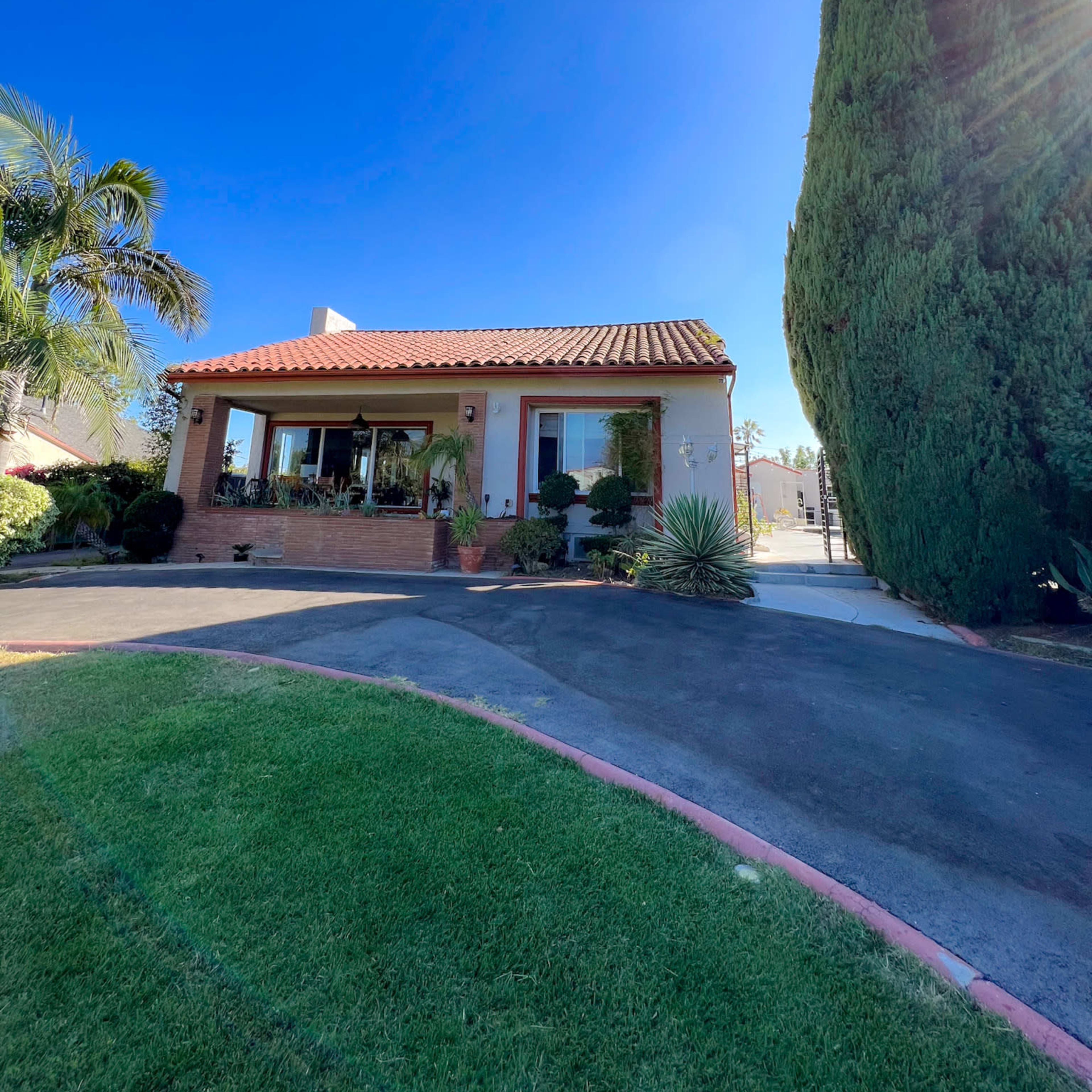 A single-story house with a red-tiled roof is situated beside a large, tall tree under a clear blue sky.