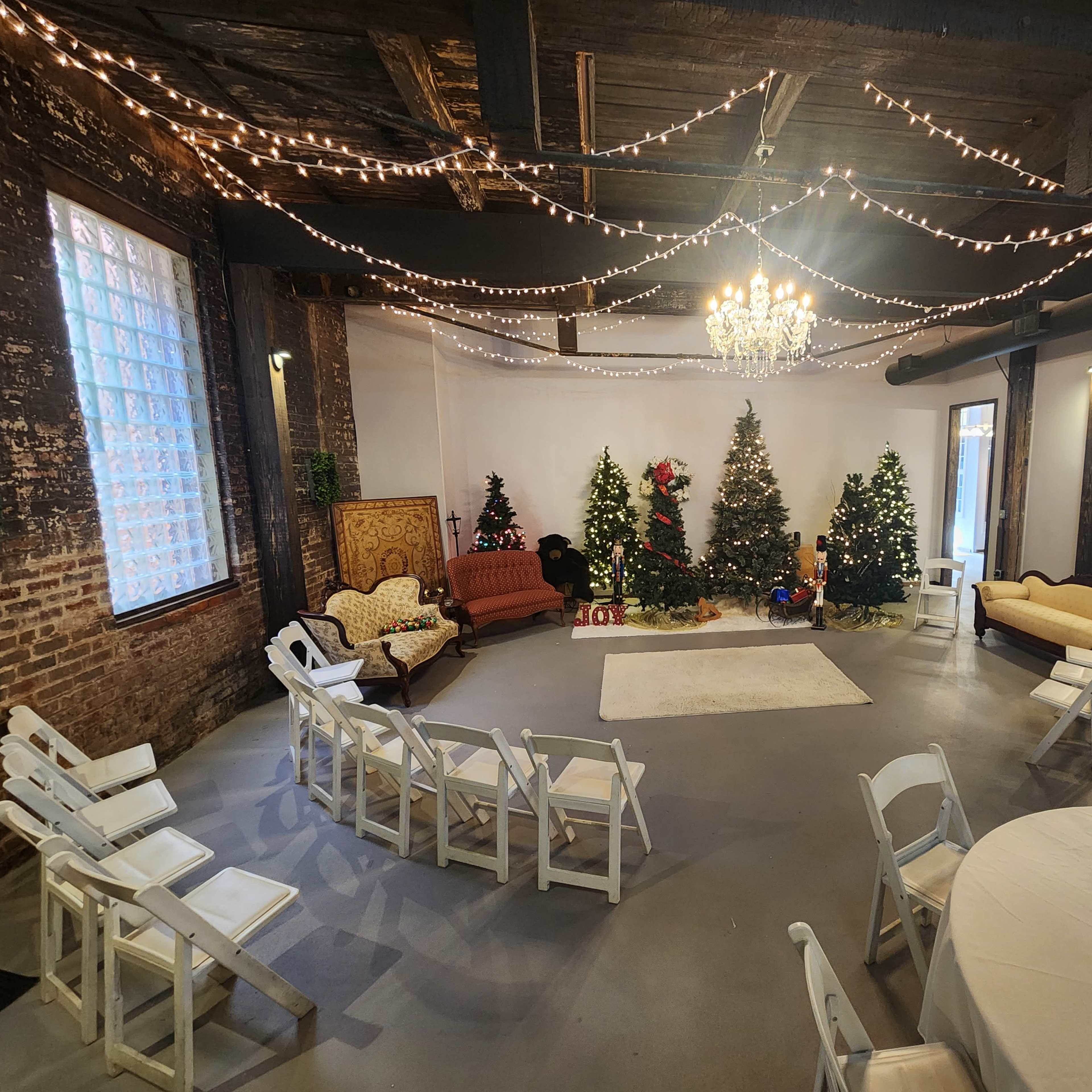 A decorated indoor event space featuring two Christmas trees, festive lighting, chairs arranged in a semicircle, and a central white carpet.