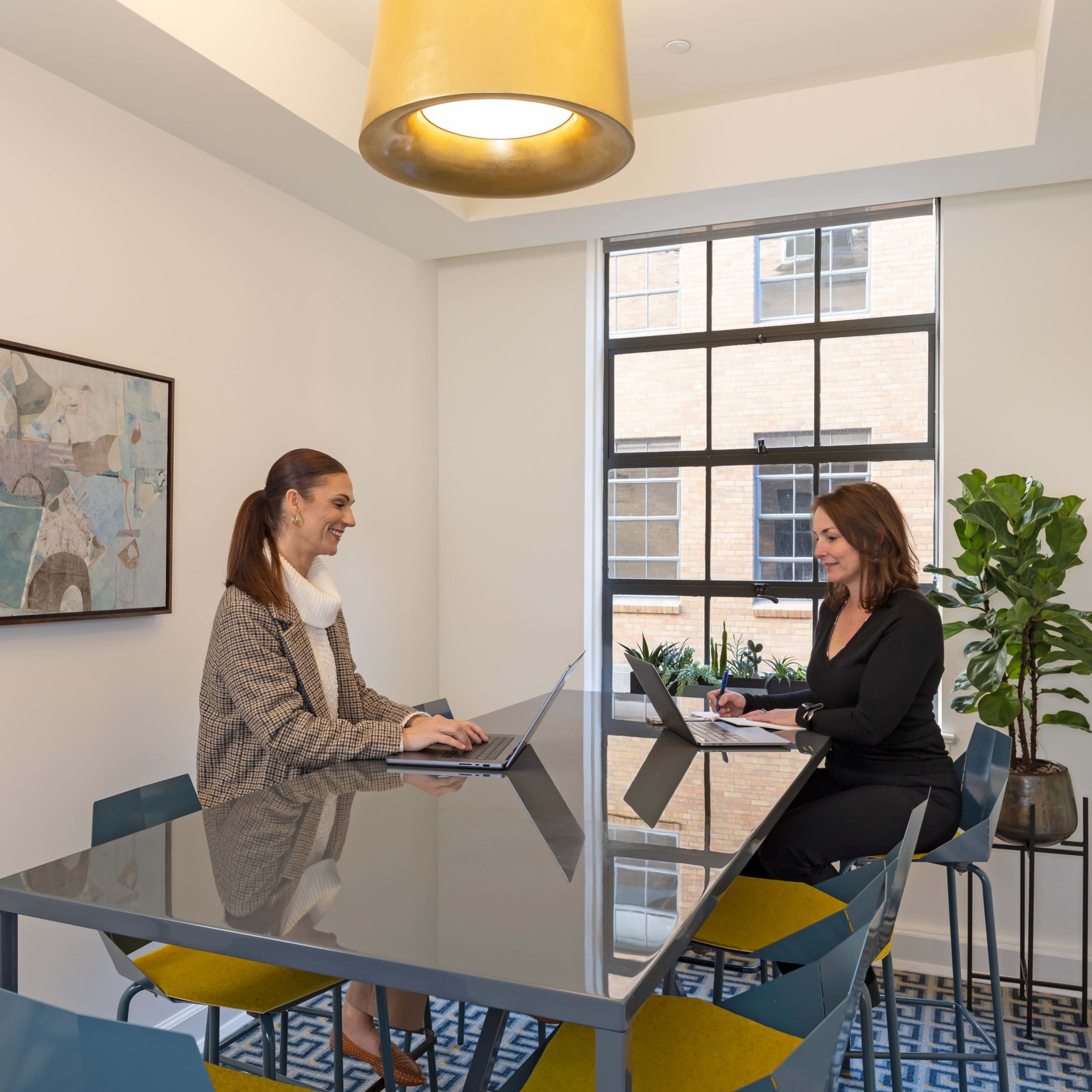 Two women are seated at a modern table in a well-lit office space, engaging in a discussion while using laptops.