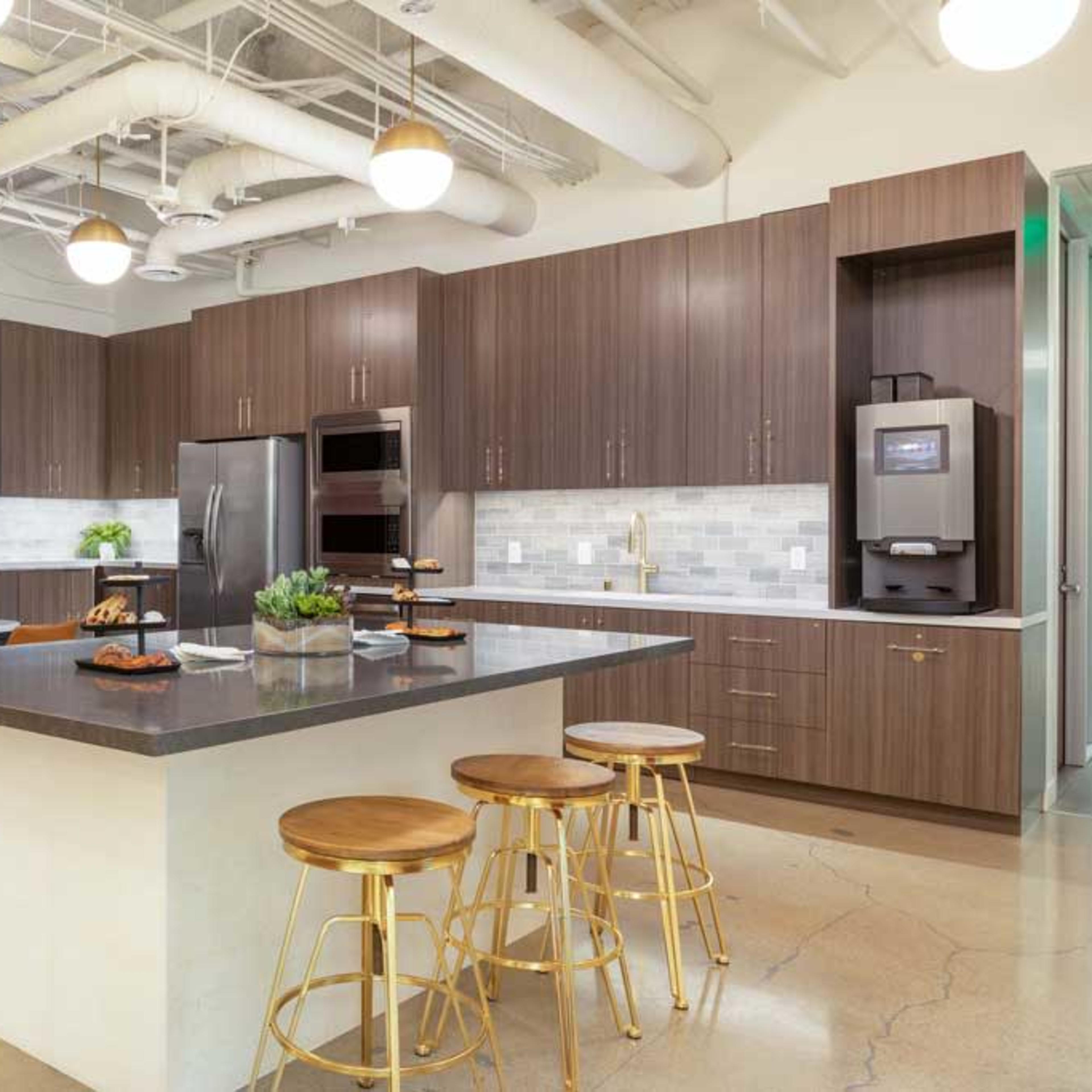A modern kitchen with dark wooden cabinetry, a central island, and four golden barstools, illuminated by hanging light fixtures.