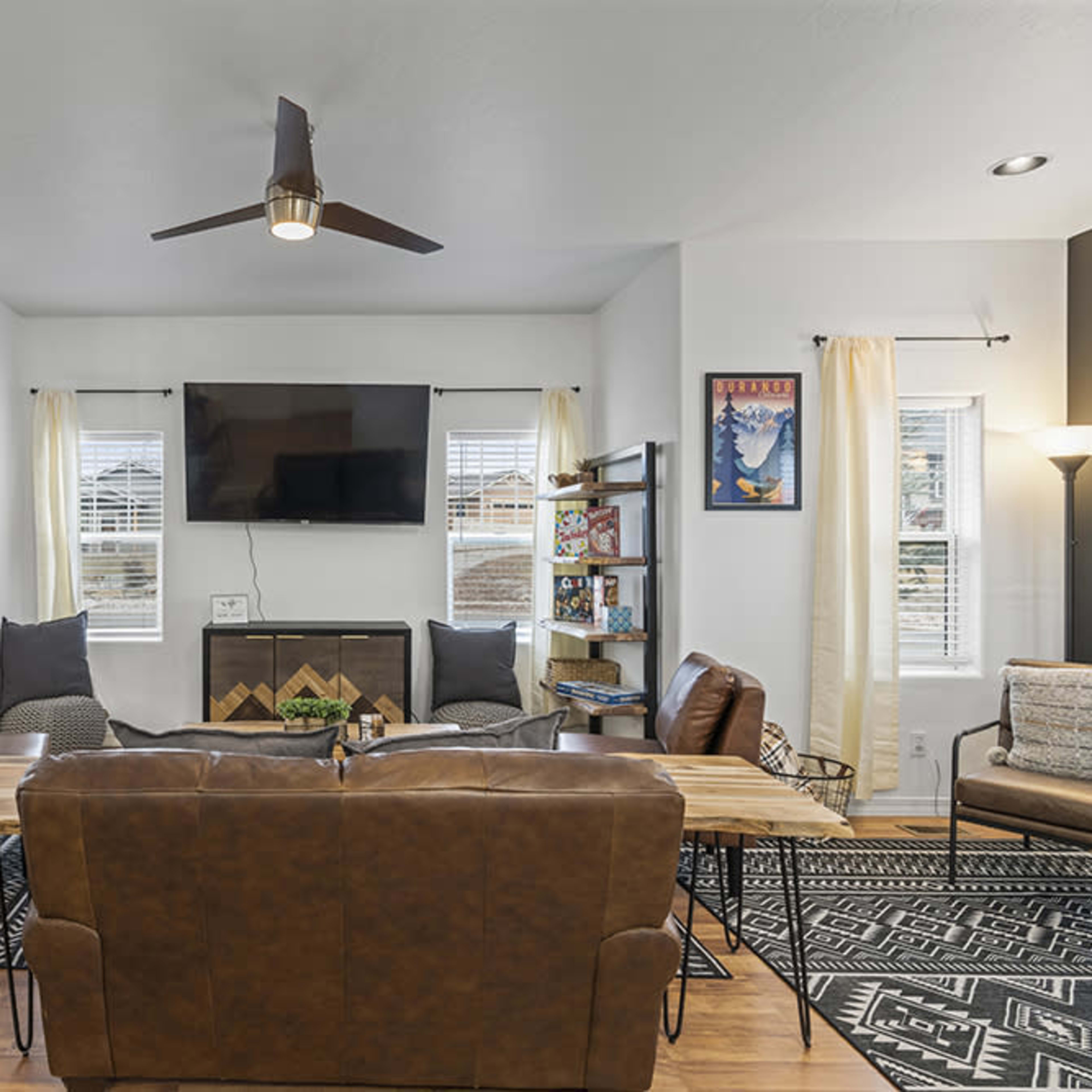 A modern living area with a flat-screen TV mounted above a fireplace, surrounded by brown leather couches, a wooden coffee table, and a dining area with black metal chairs in the background.