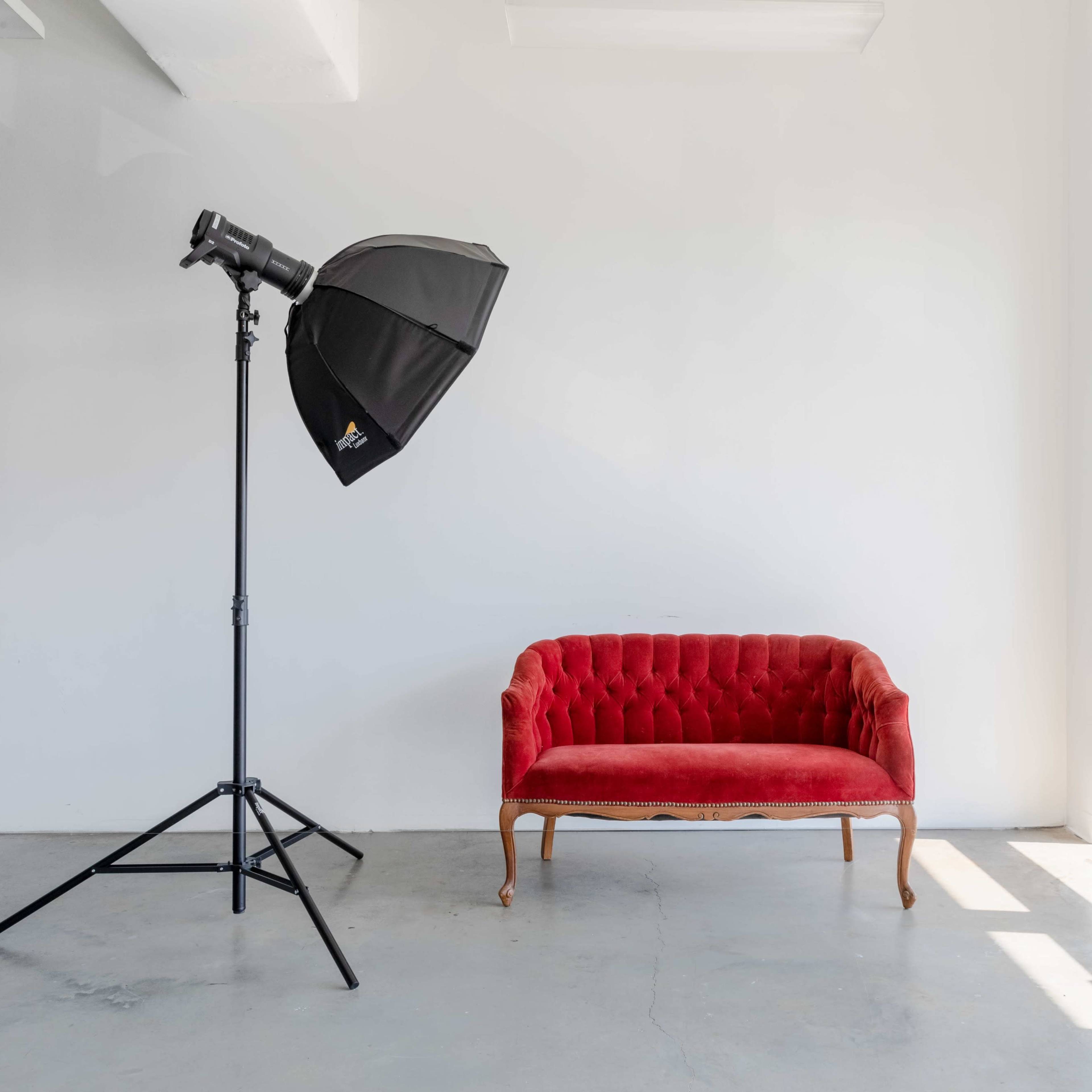 A red velvet sofa is positioned next to a large window, with a black photography umbrella reflector on a stand nearby.