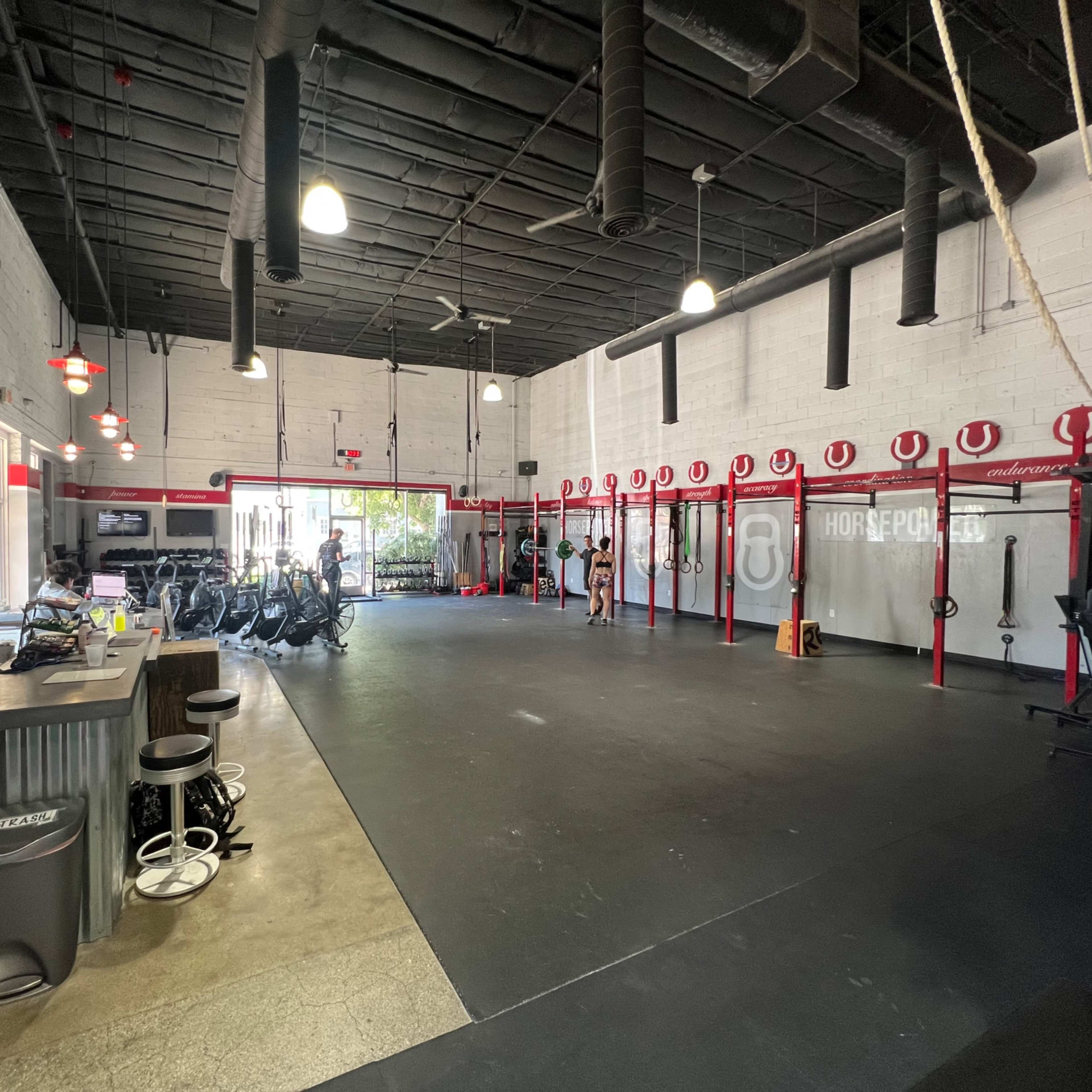 An interior view of a gym with exercise equipment arranged against the walls and a few people engaged in workouts.