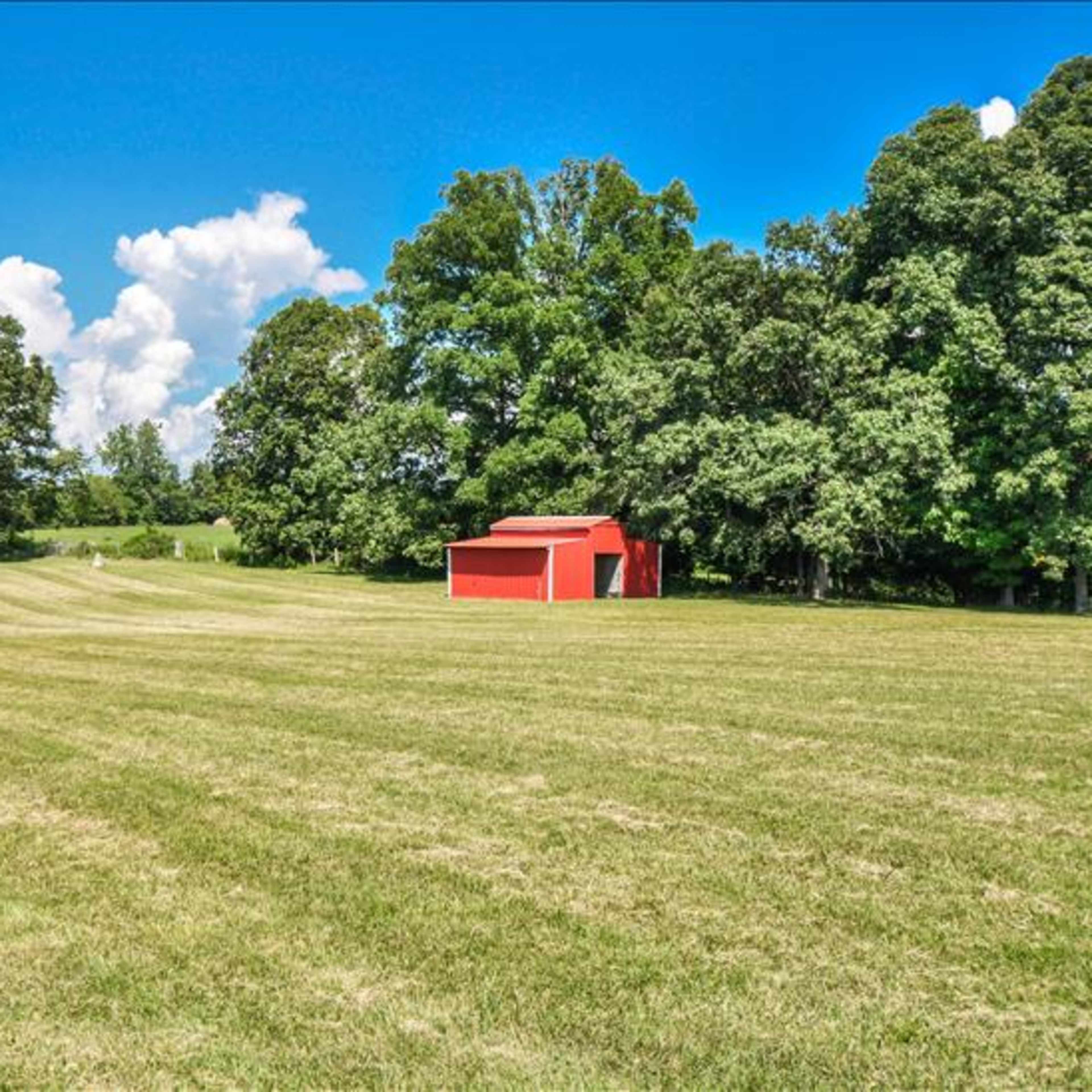 A red barn stands in a grassy field surrounded by trees under a bright blue sky.