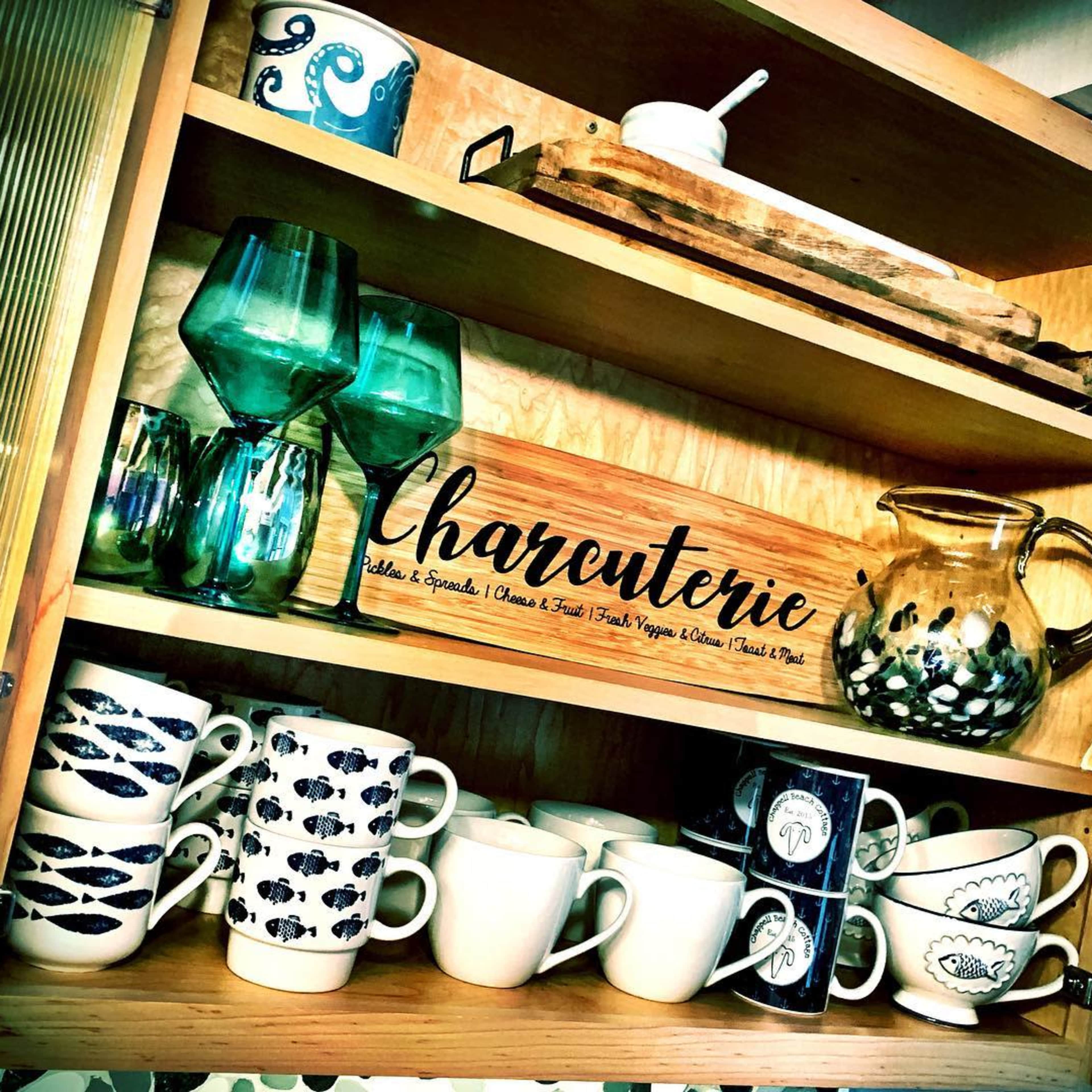 A wooden cabinet with shelves containing a variety of glassware, ceramic cups, and a decorative sign labeled "Charcuterie."