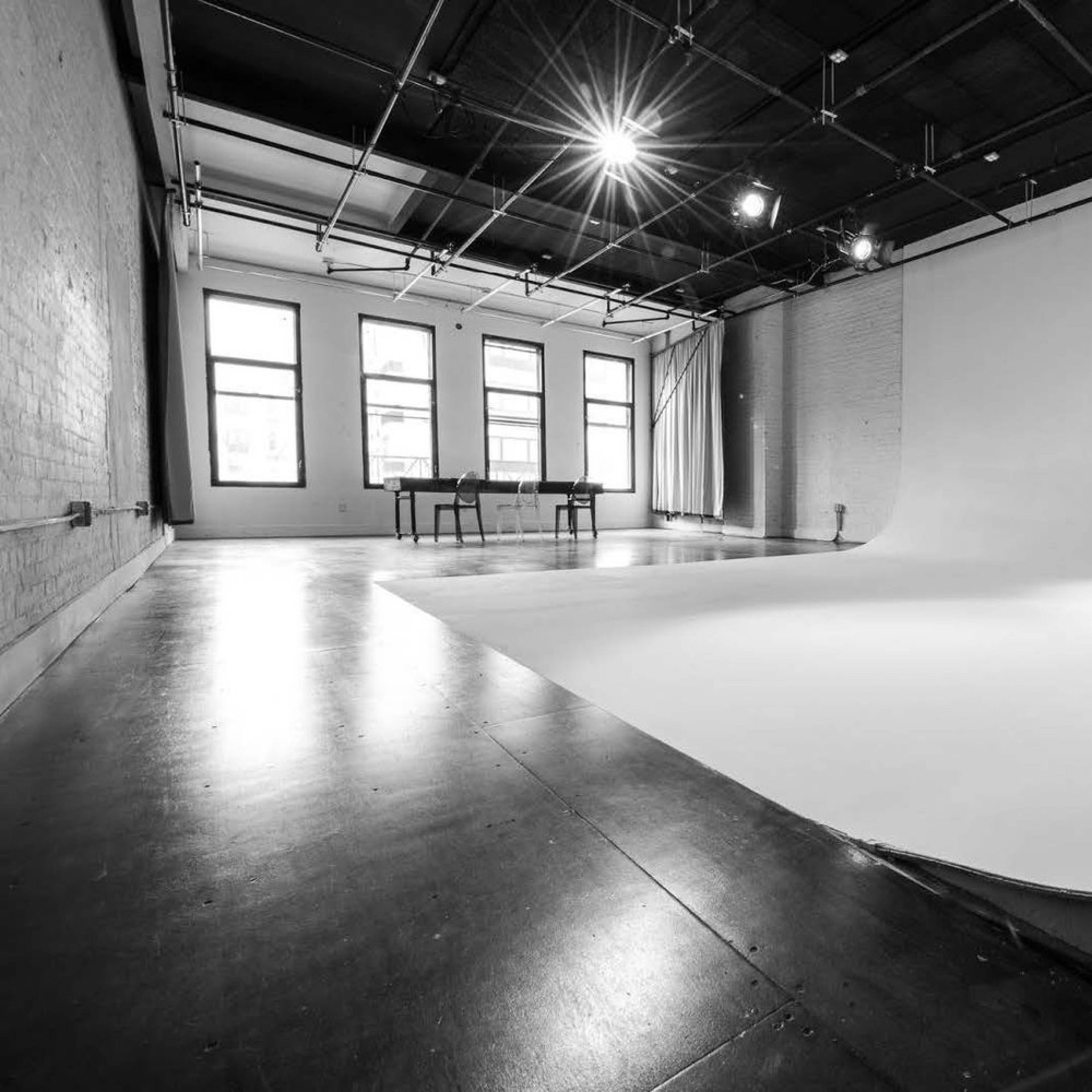A spacious photography studio features a seamless white backdrop, large windows, and a simple table in the background.