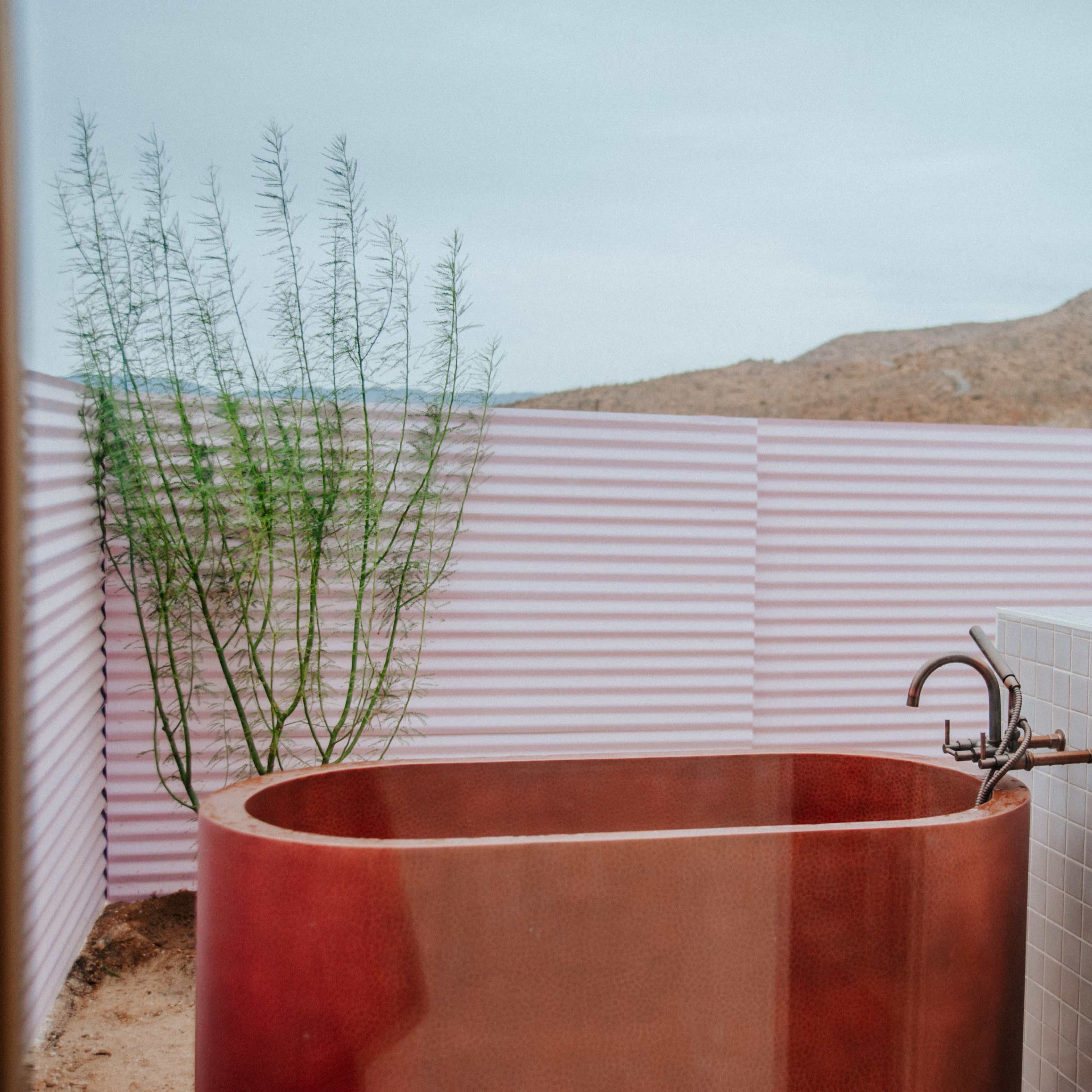 A stylish outdoor bathtub made of metal, surrounded by a pink corrugated fence, with a desert landscape in the background.