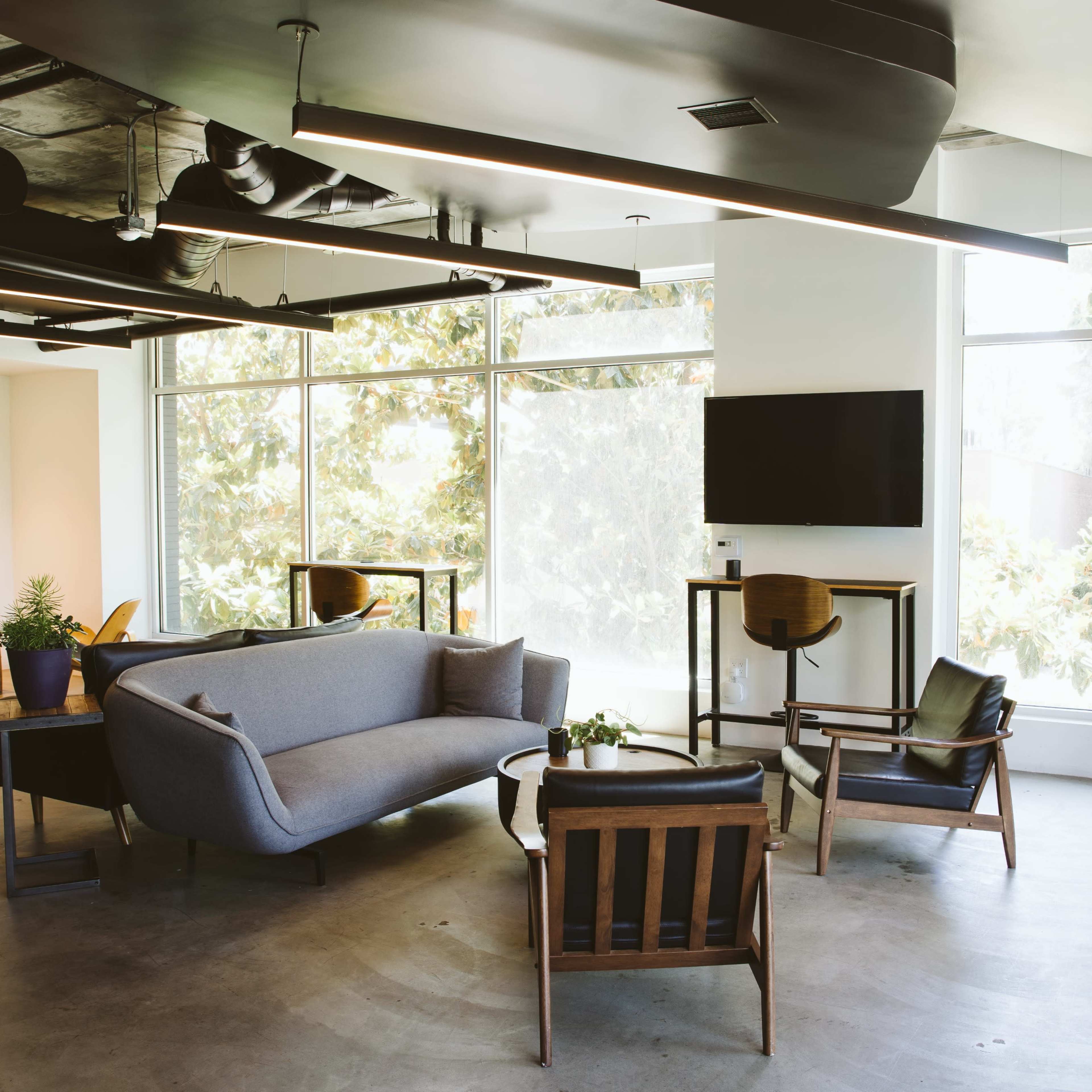 A modern lounge area with large windows, contemporary furniture, and a TV mounted on the wall.