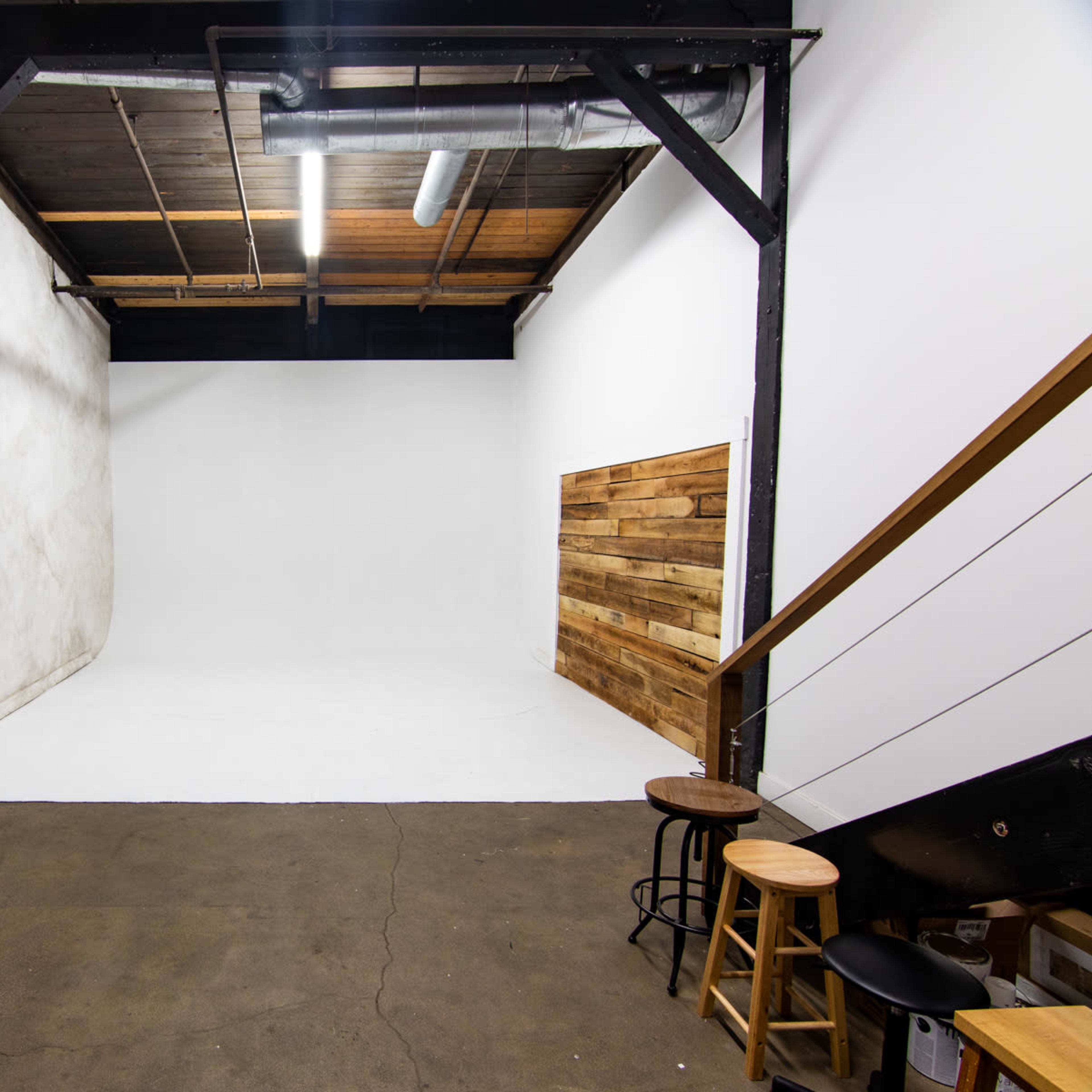 A spacious photography studio with a white backdrop, wooden accent wall, and two light stands beside a staircase.