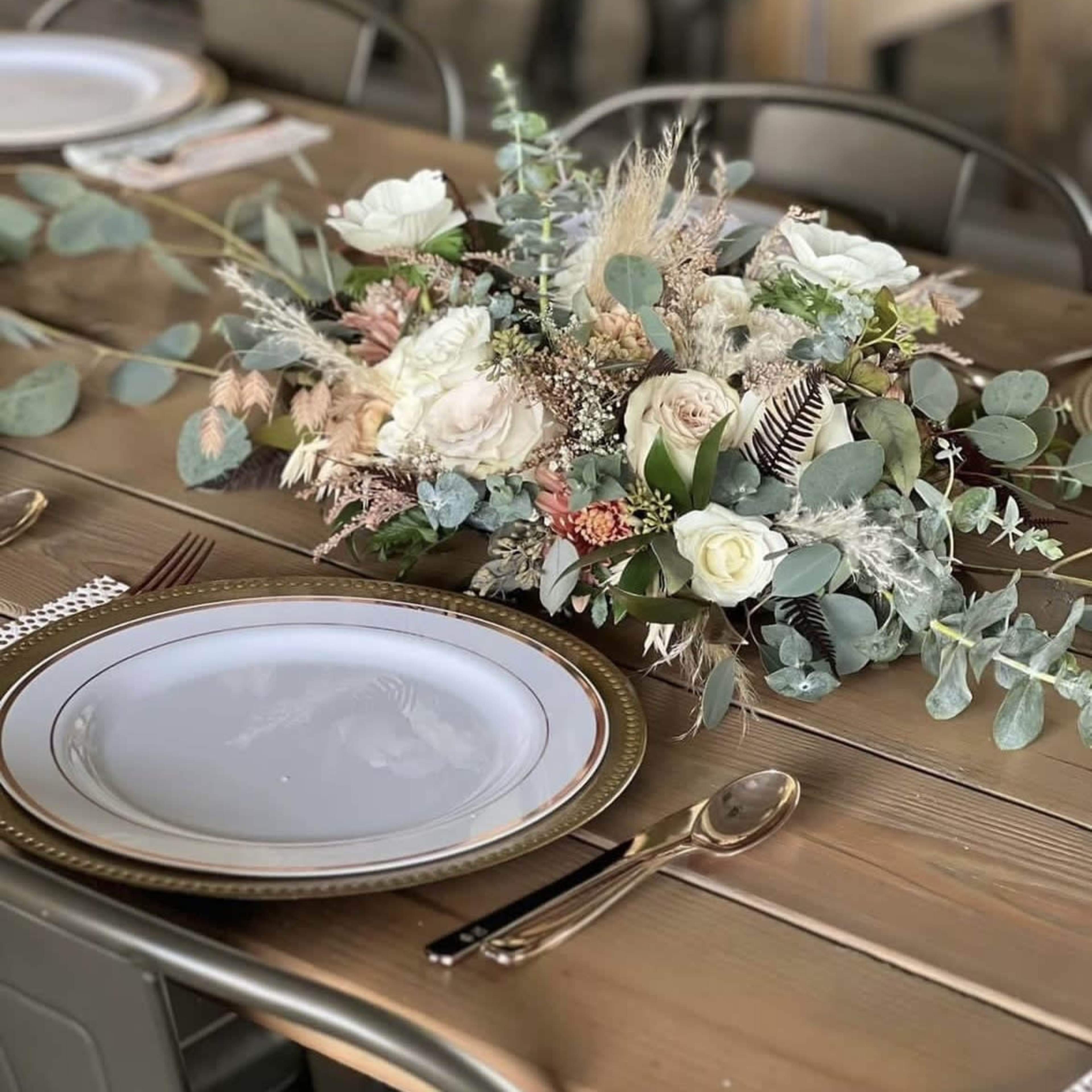 A floral centerpiece made of various dried and fresh flowers is placed on a wooden dining table set with white plates and shiny cutlery.