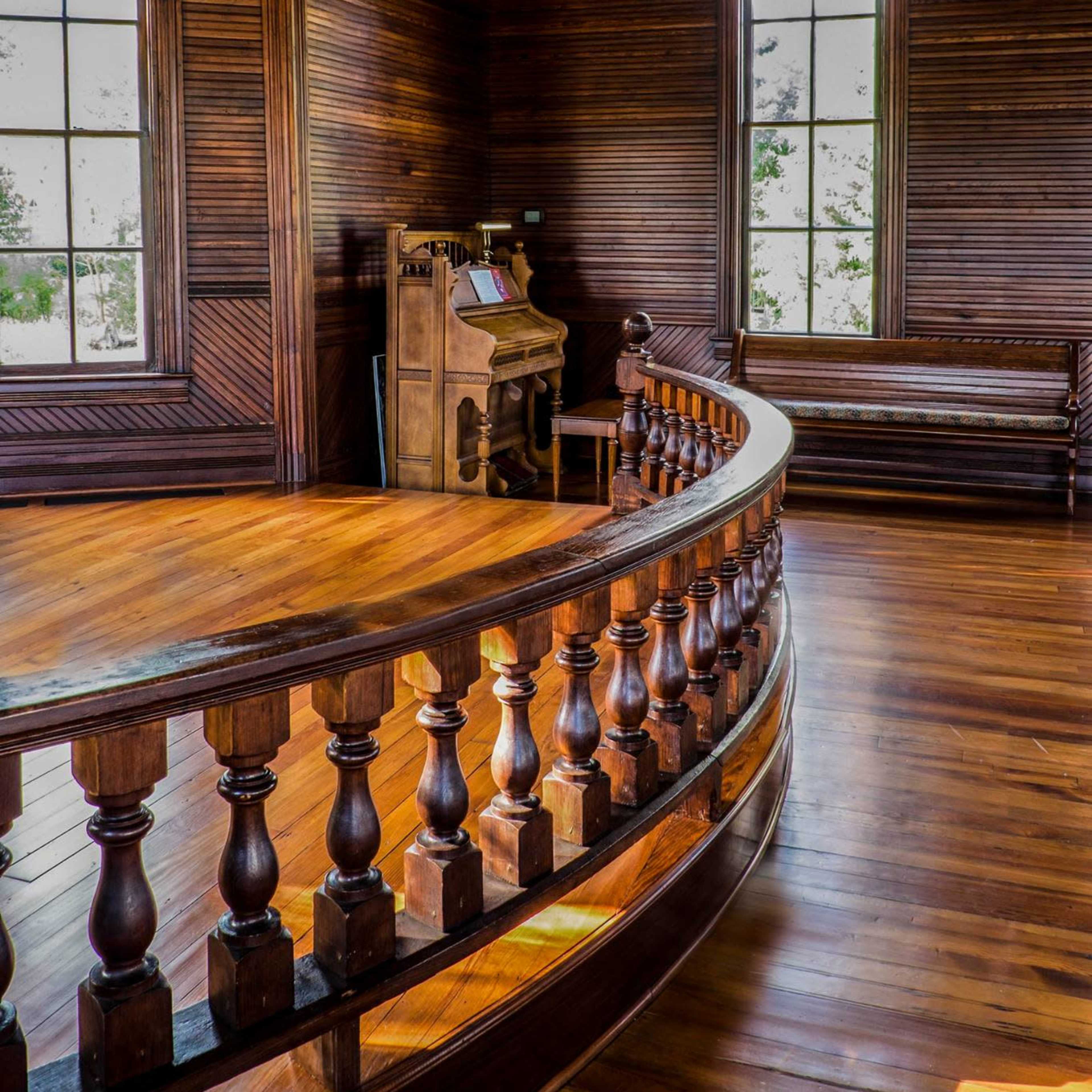 A wooden interior of a room featuring large windows, a curved railing, and an antique organ placed against the wall.