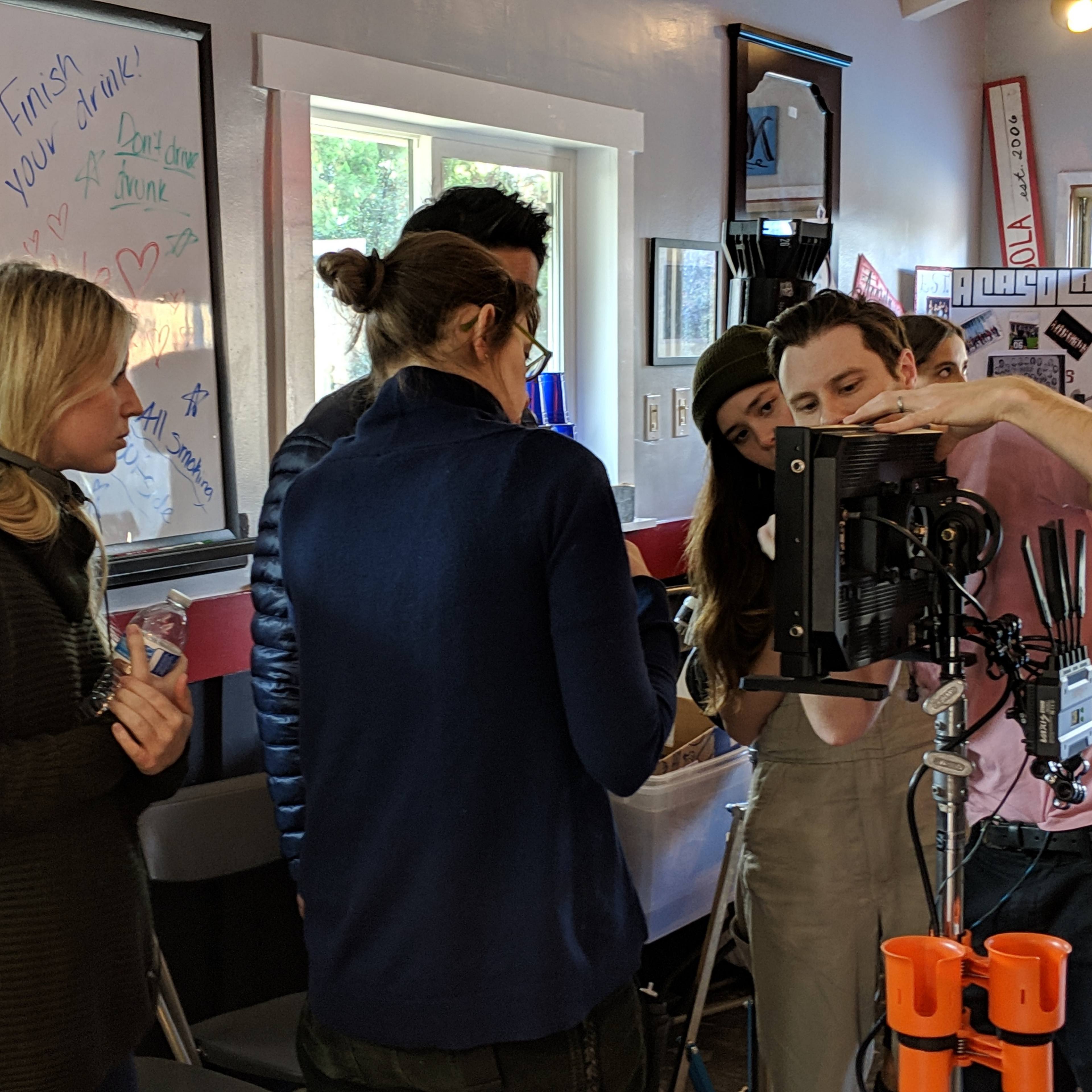 A group of four people inspects a camera setup while another person observes in a brightly lit room decorated with notes and posters.