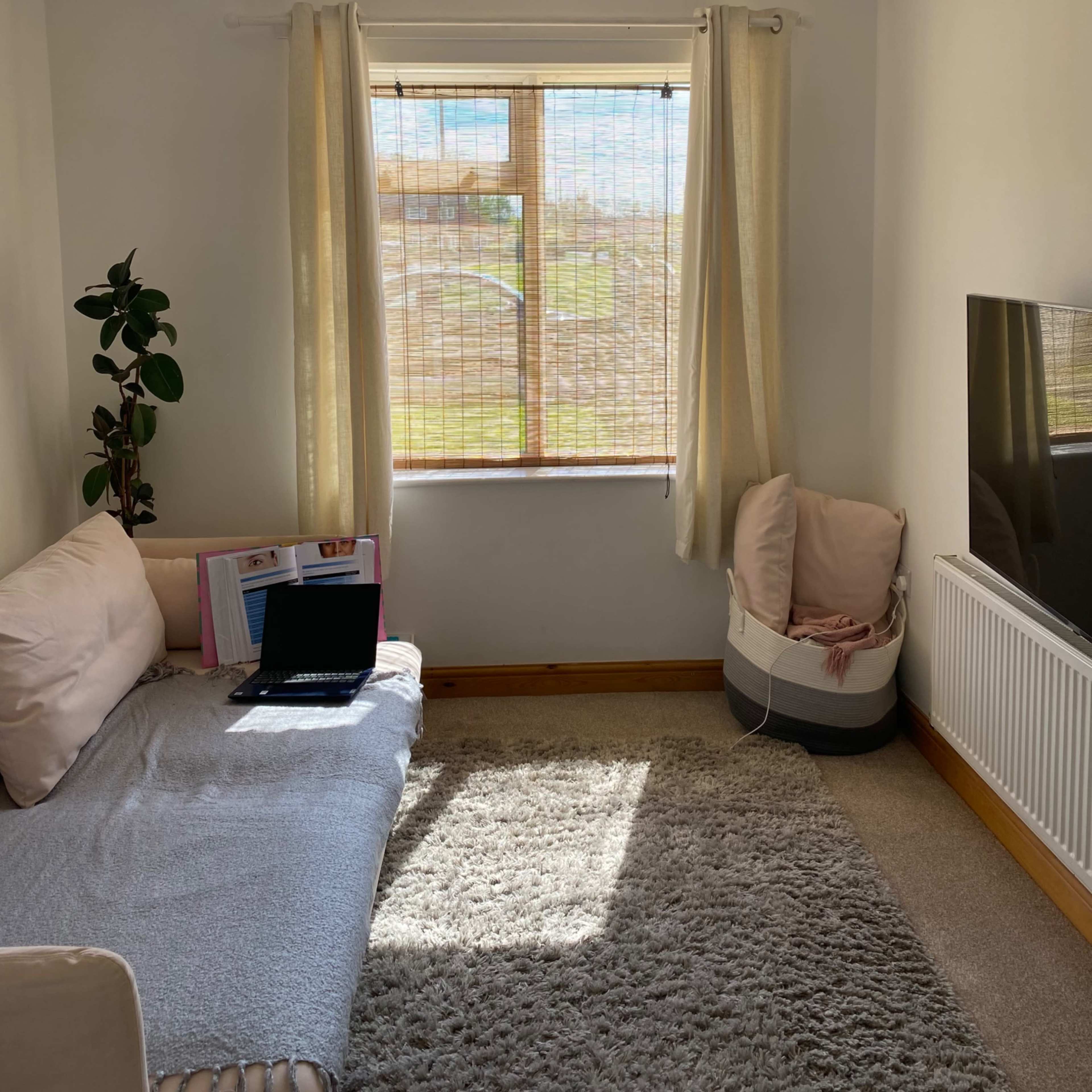 A small, well-lit room features a beige couch, a laptop on a soft throw, and a large window with light curtains, revealing a view of an outdoor area.