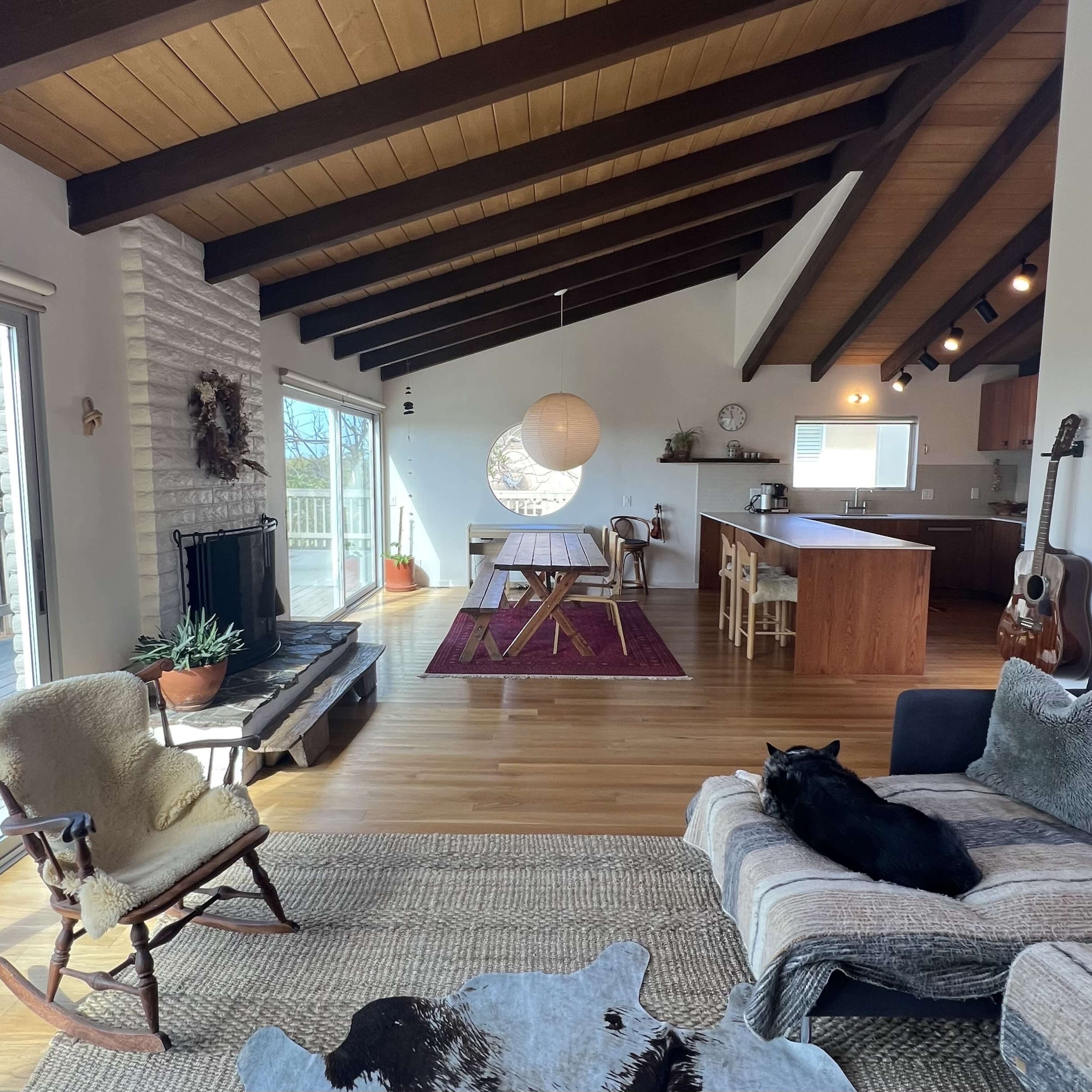 A spacious living area featuring a seating arrangement, dining area, and an open kitchen with wooden beams and large windows.