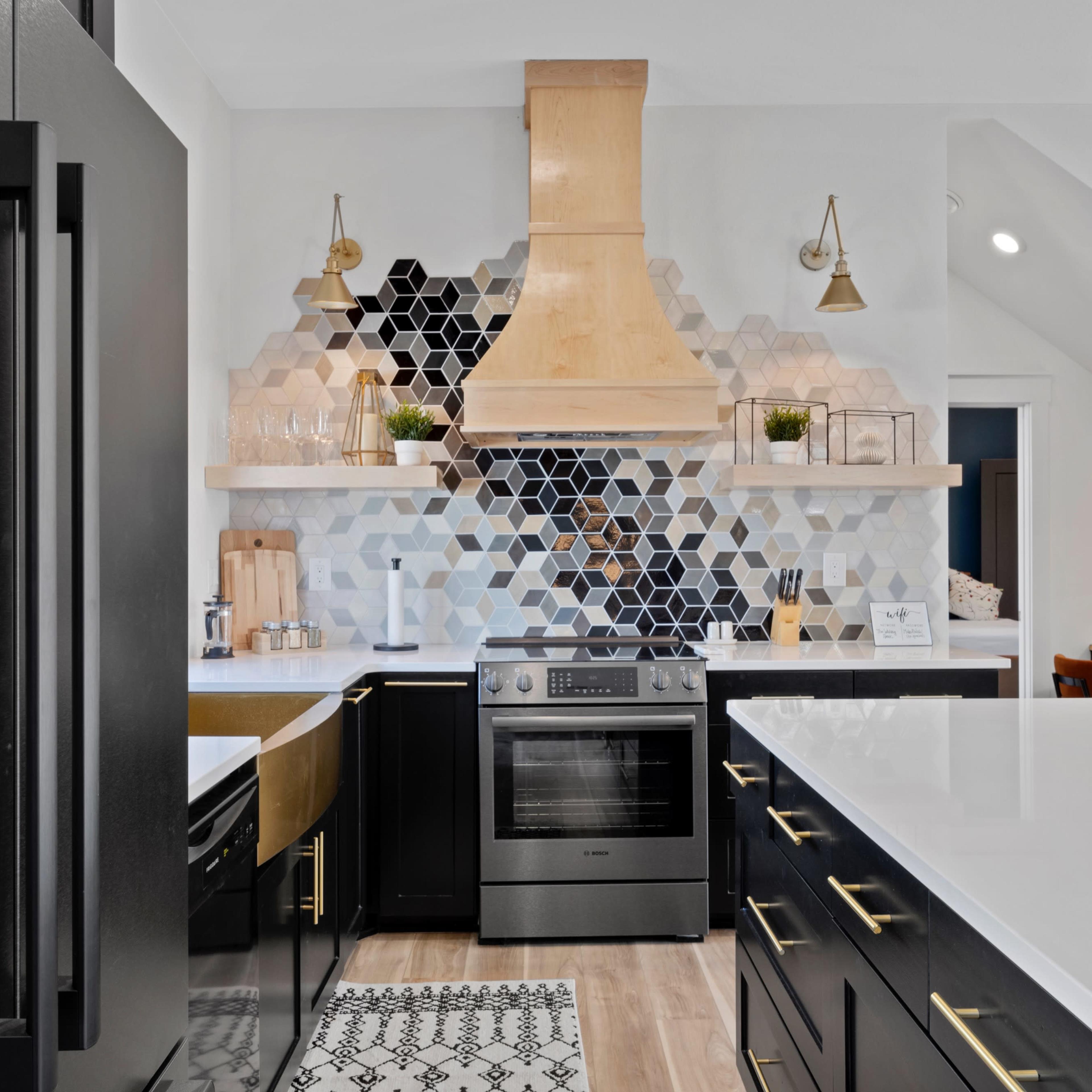 A modern kitchen features a geometric black and white tile backsplash, a wooden range hood, and sleek black cabinetry with brass hardware.