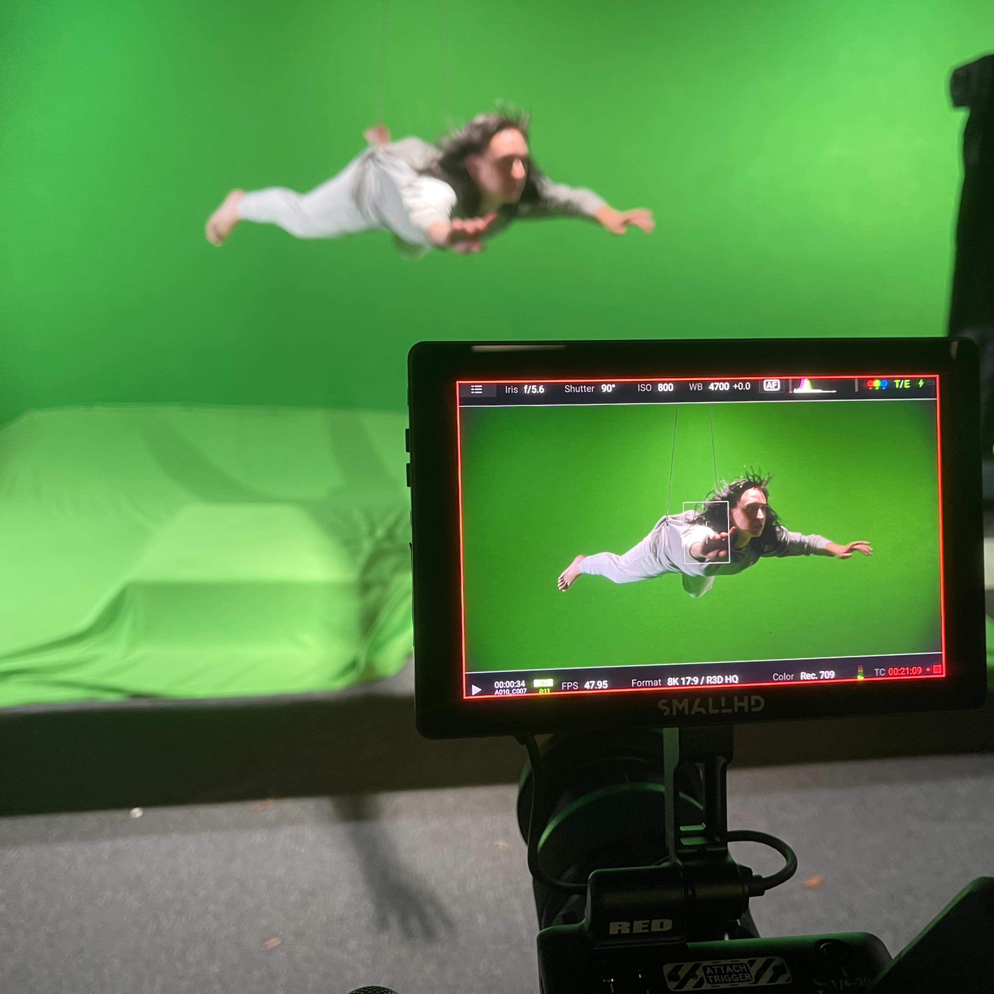 A person is suspended in mid-air in front of a green screen, while a camera monitor displays the scene.