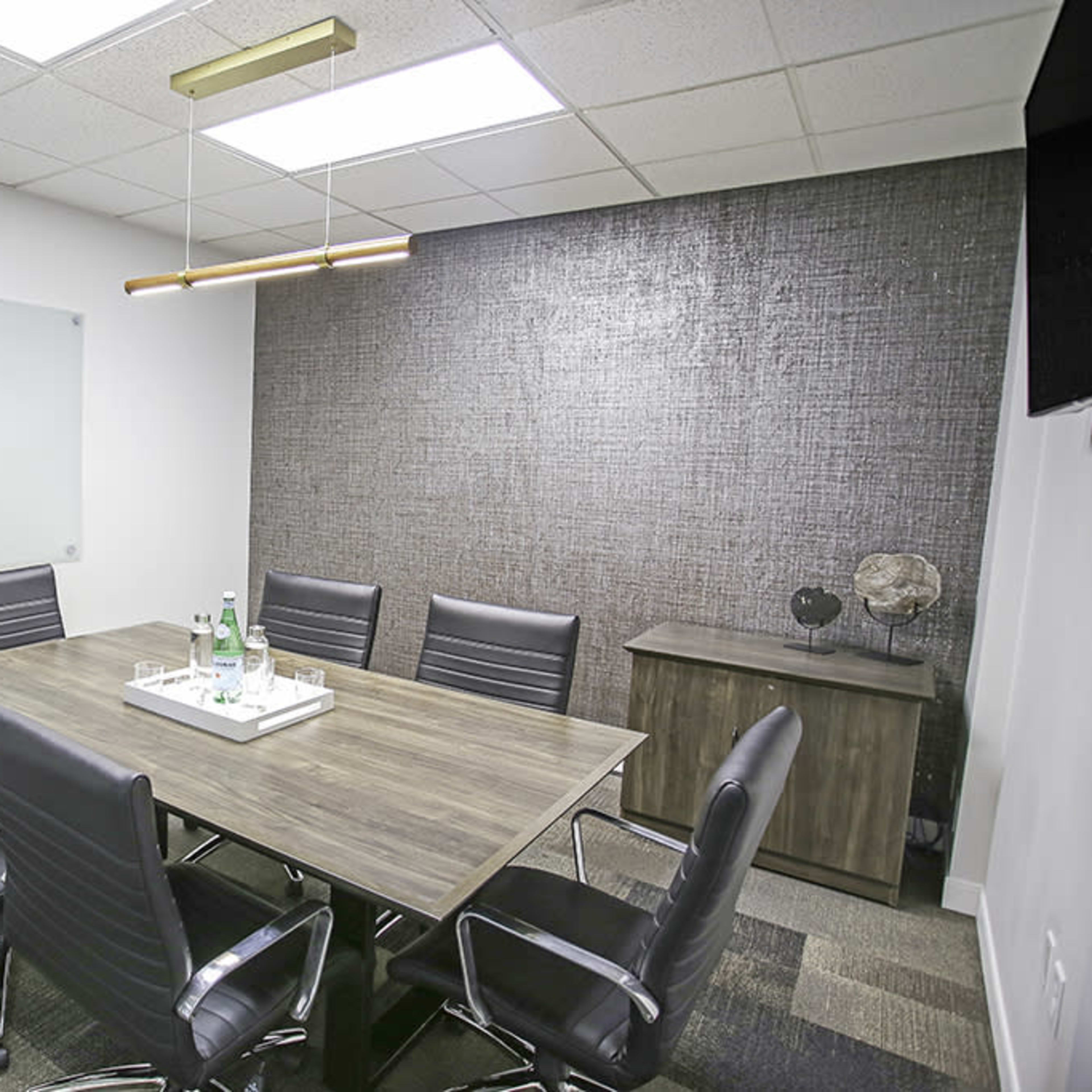A modern conference room features a rectangular wooden table surrounded by black leather chairs, with a glass whiteboard on one wall and a textured accent wall.