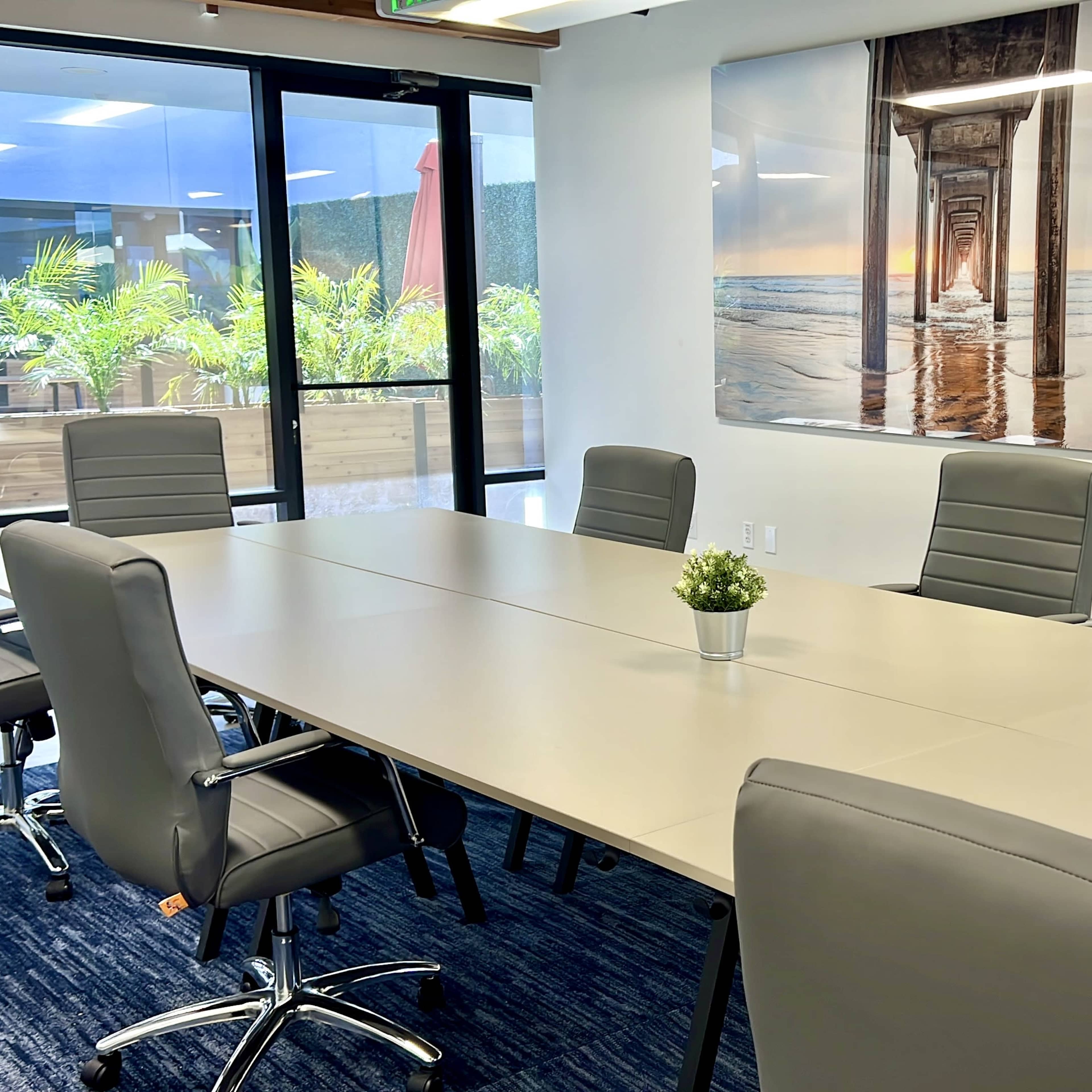 A modern conference room features a large rectangular table surrounded by ergonomic chairs, with a beach-themed artwork on the wall and large glass doors leading to an outdoor area.