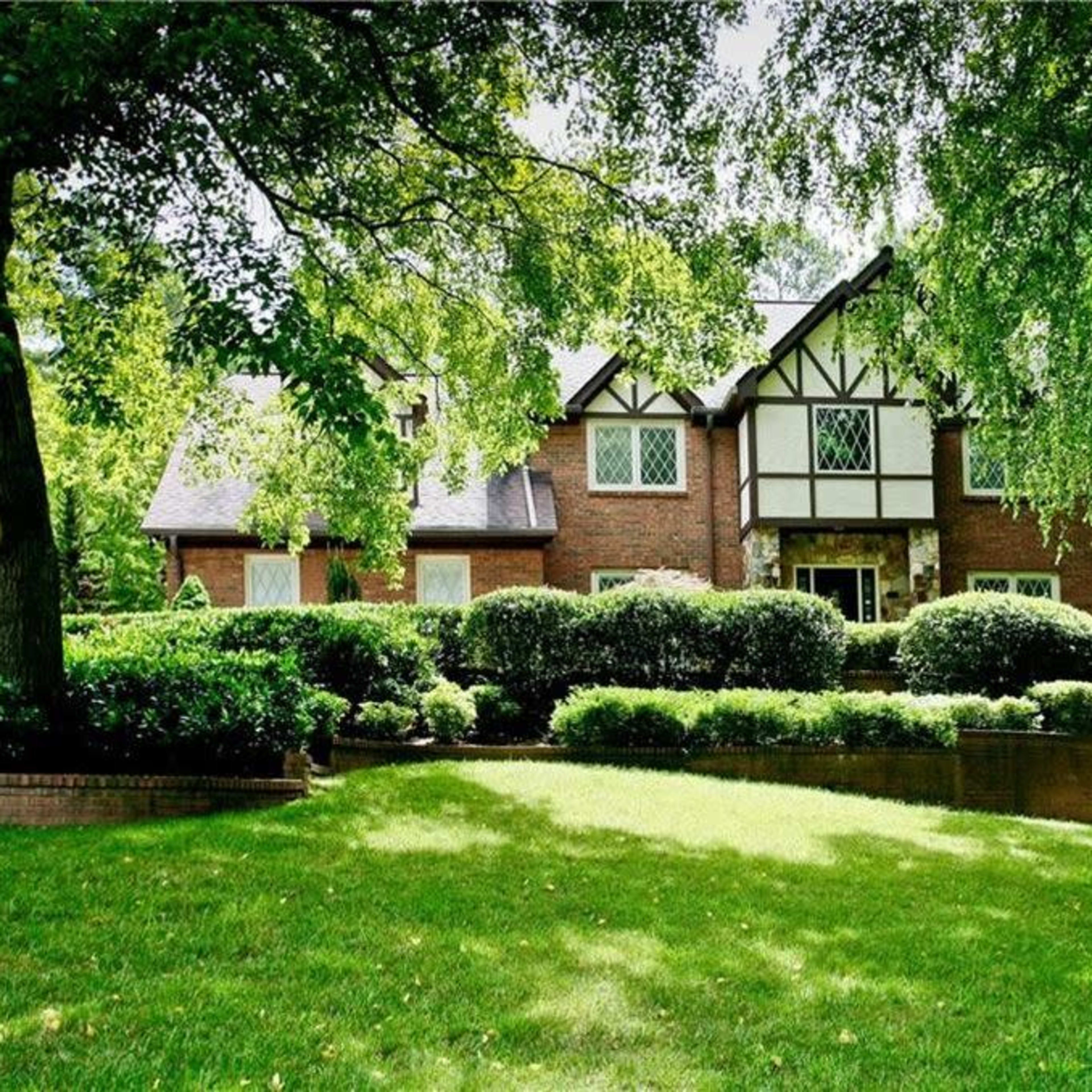 A large brick house with Tudor-style architecture is set on a lush green lawn surrounded by dense trees and neatly trimmed shrubs.