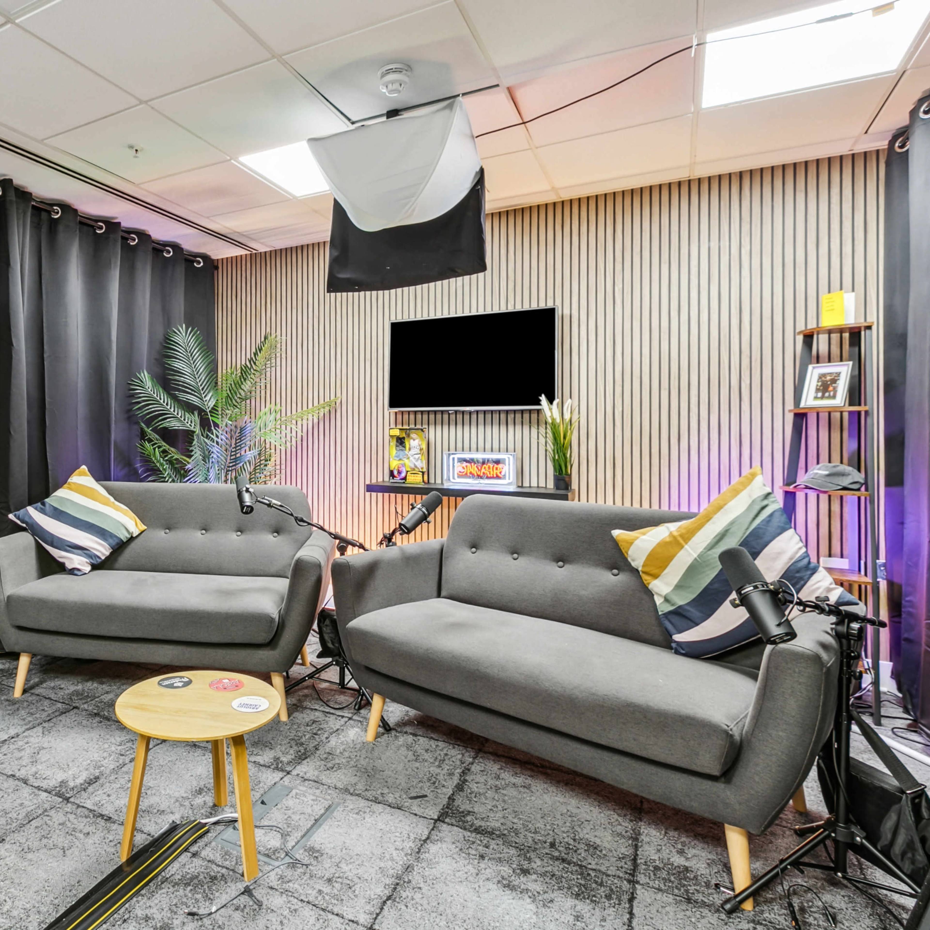 A studio space with two gray sofas, a wooden coffee table, and photography equipment in front of a backdrop of black curtains and a wooden-paneled wall.