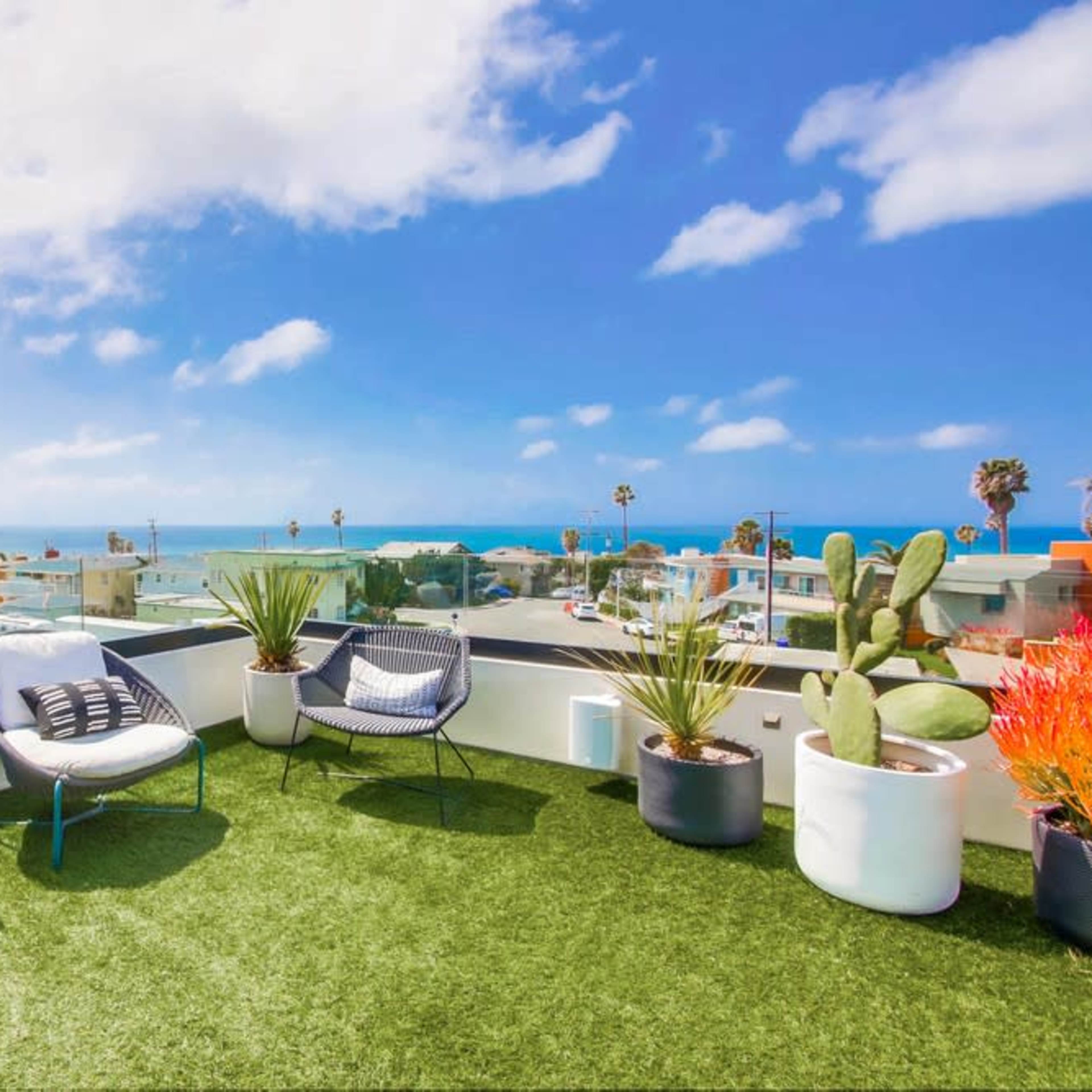 A rooftop terrace features four chairs, potted plants, and a view of the ocean under a blue sky.