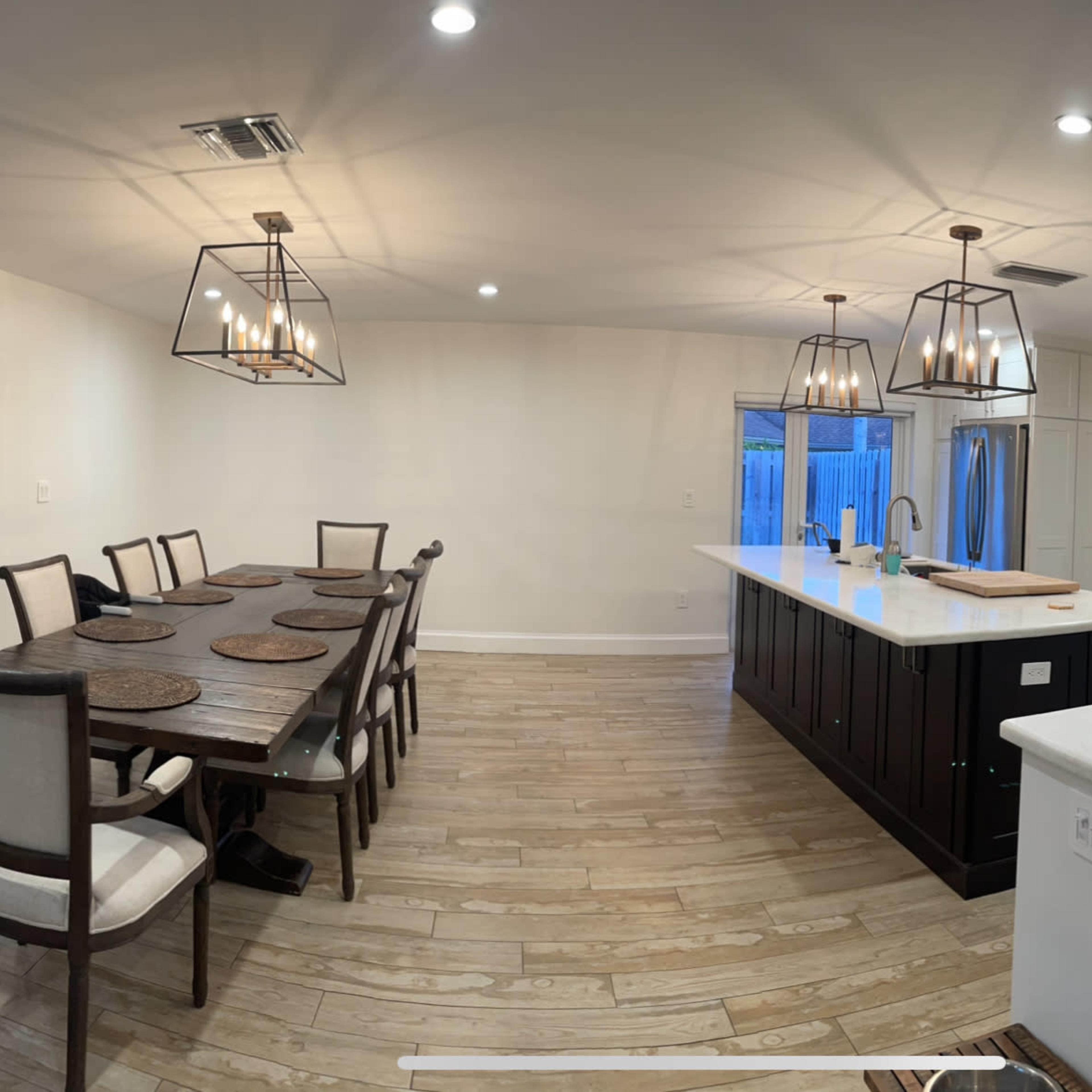 A modern kitchen and dining area featuring a long dining table with chairs, an island with seating, and large windows facing an outdoor space.