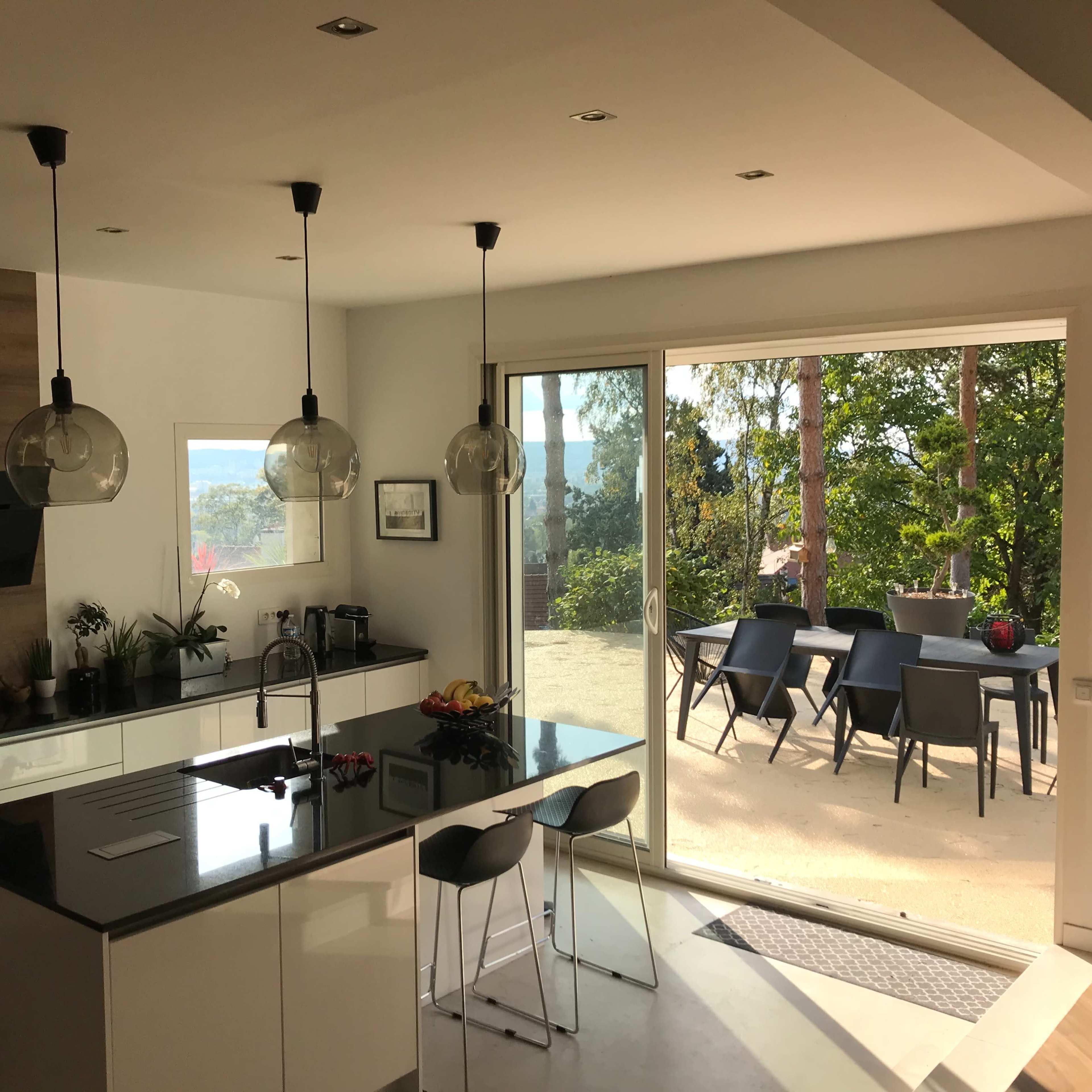 A modern kitchen features a central island with three stools, large windows opening to a patio area with outdoor seating and views of greenery.