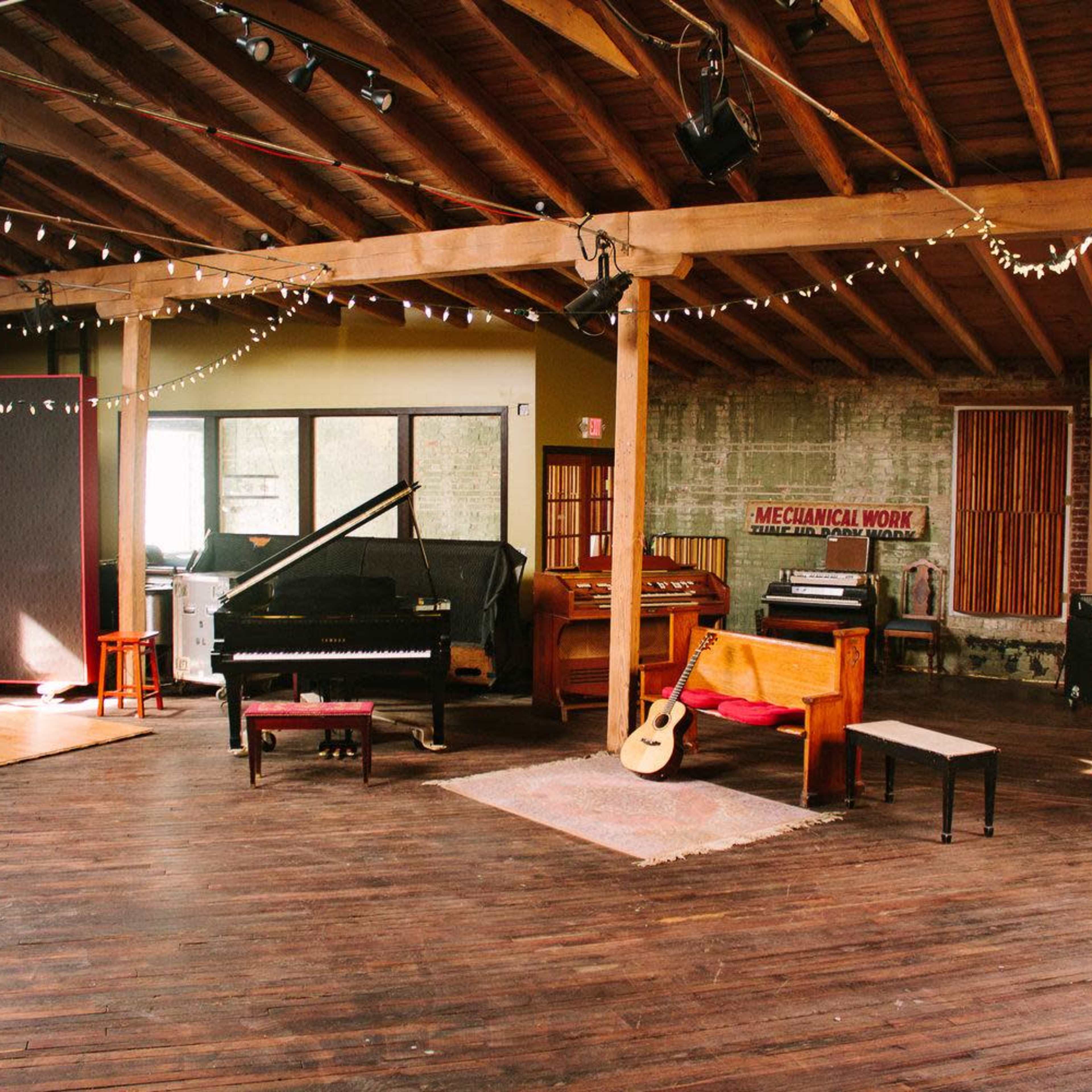 A spacious music studio with a grand piano, various instruments, and warm wooden floors under ambient string lights.