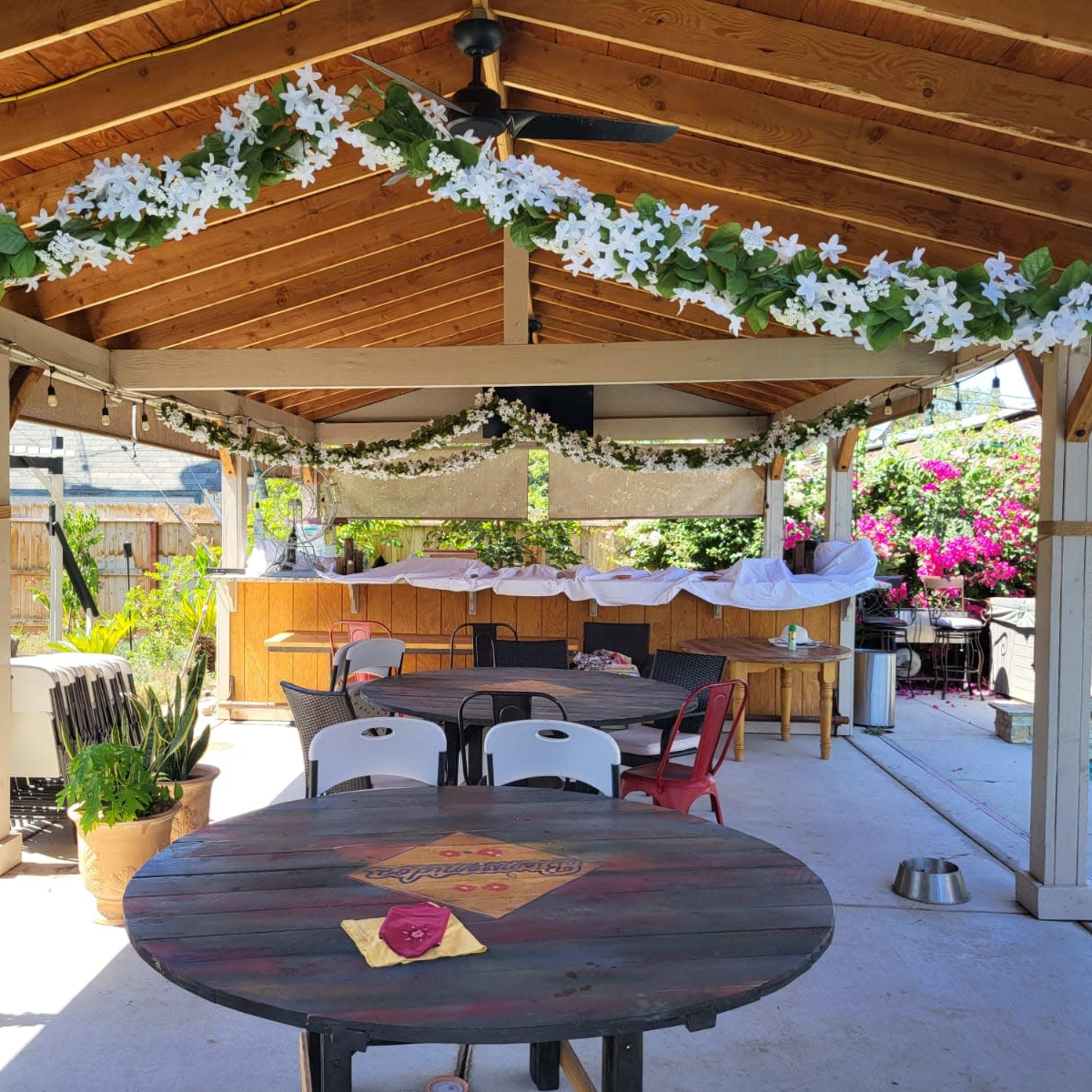 A covered outdoor space features a wooden ceiling with floral decorations, tables, and a pool in the background.