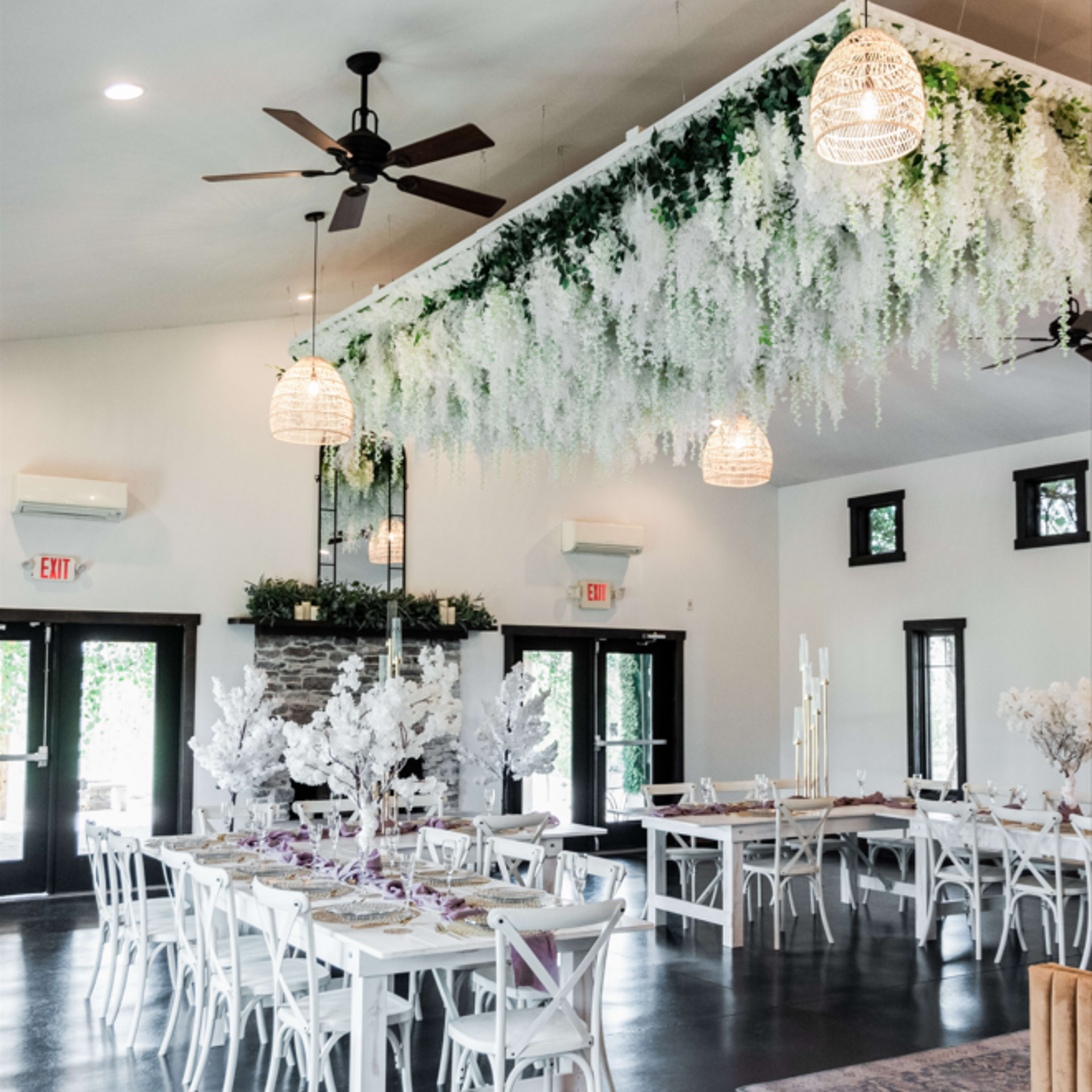 The interior of a spacious event venue features tables with white chairs, adorned with floral decorations, and a ceiling installation of greenery and hanging pendant lights.