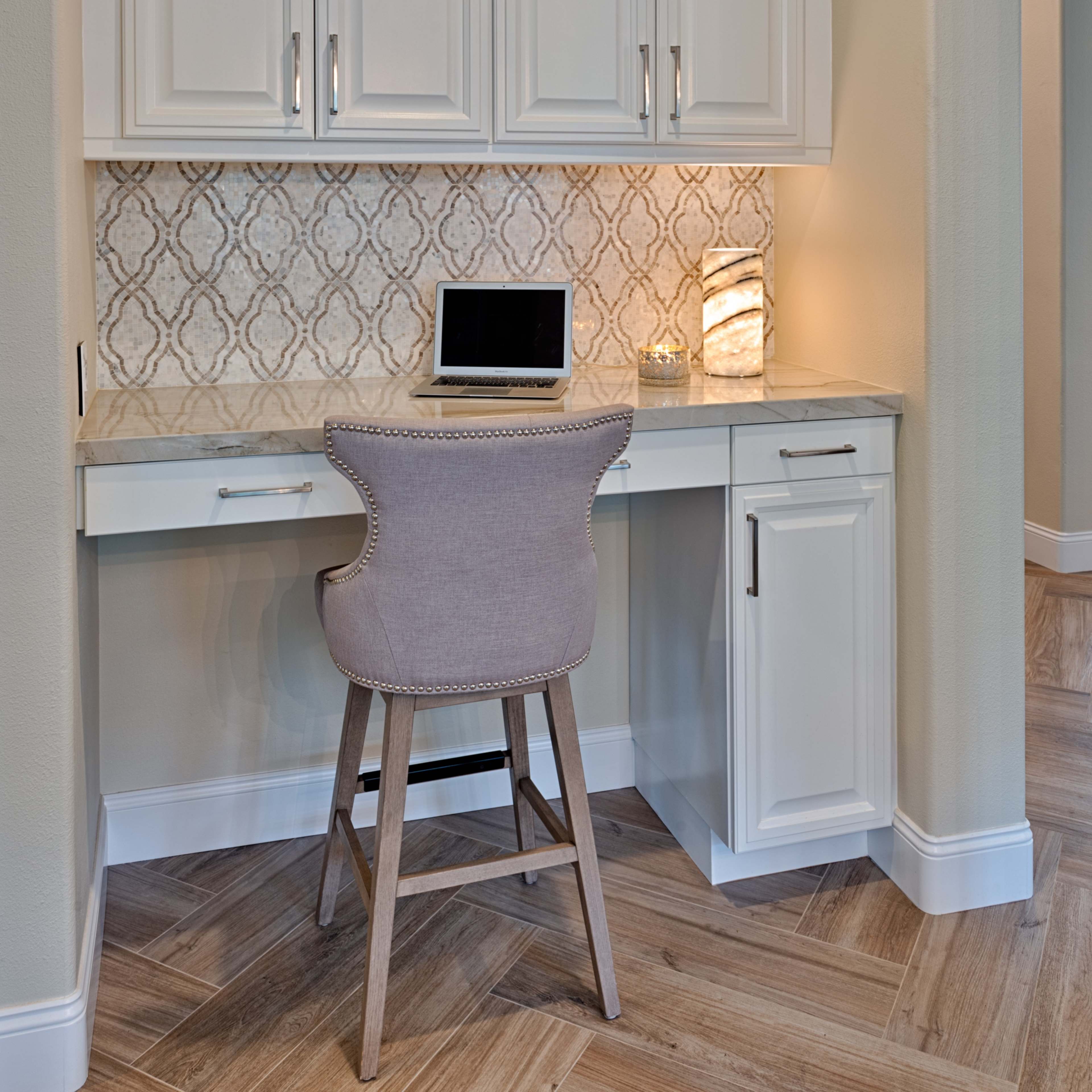 A small home office nook featuring a white desk, a light-colored chair, a laptop on the countertop, and patterned backsplash beneath white cabinets.