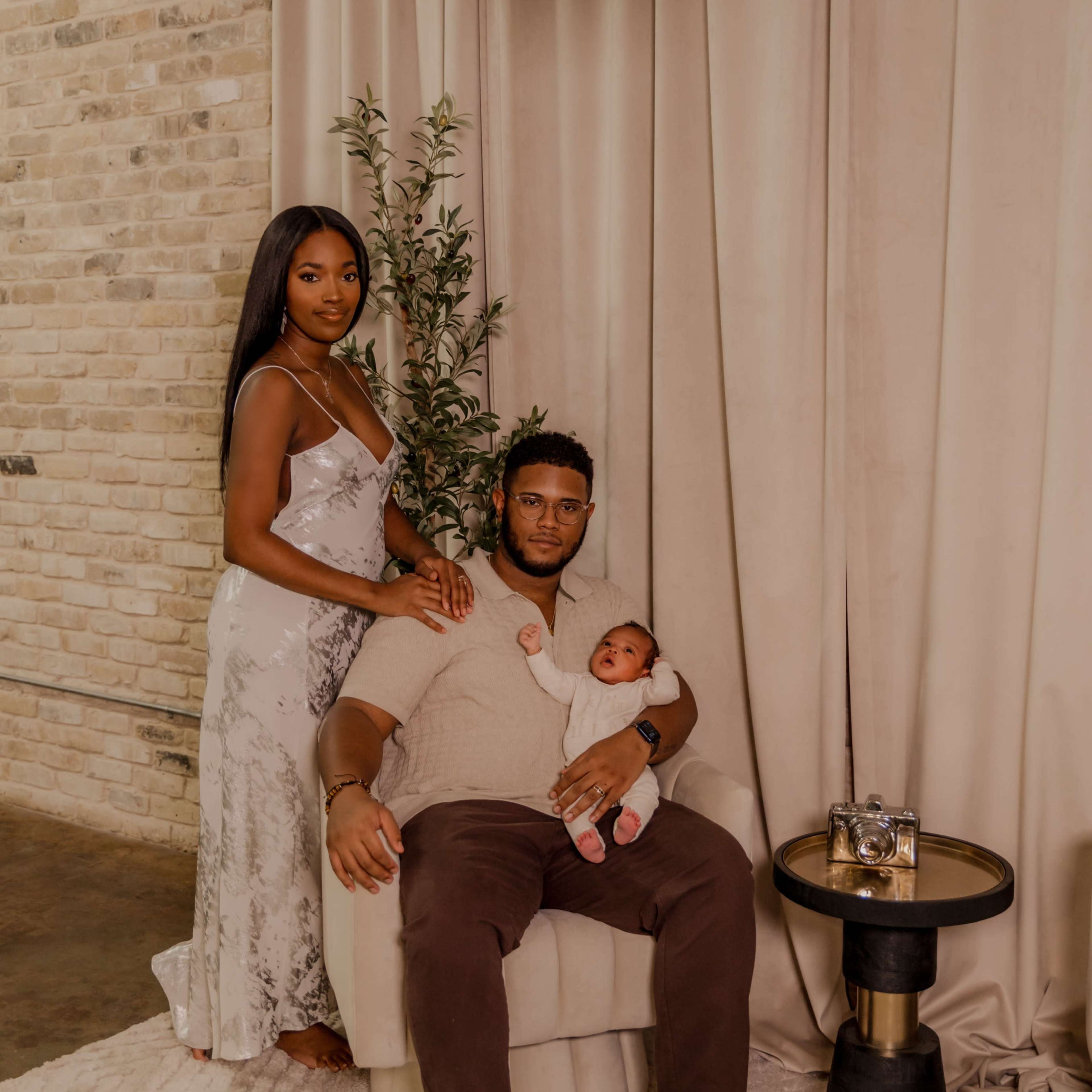 A woman stands next to a man seated in a chair, holding a baby, in a softly lit room with curtains and a plant in the background.