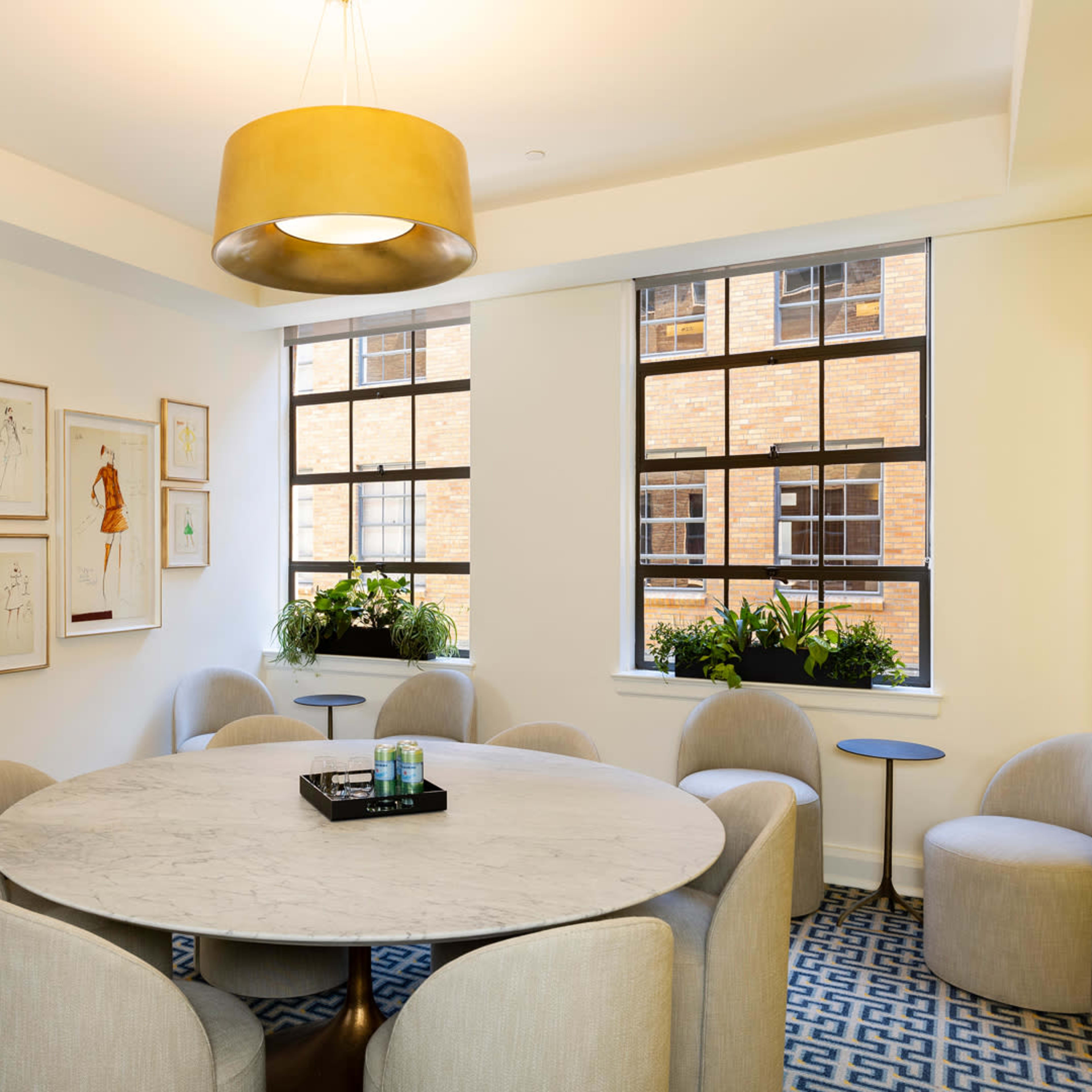 A circular marble table surrounded by upholstered chairs is situated in a well-lit room with large windows and framed artwork on the walls.